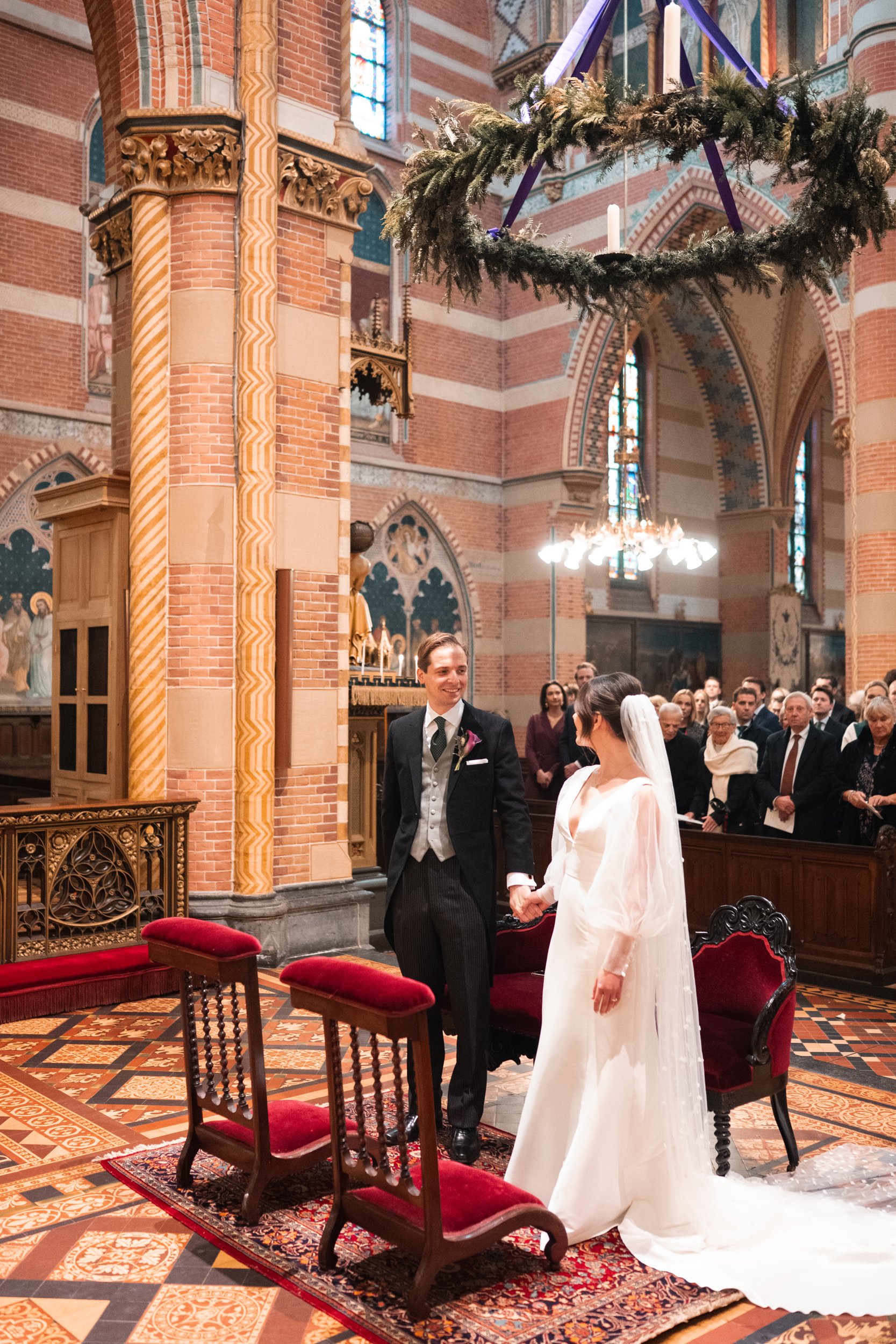 A bride and groom exchanging vows at their wedding ceremony inside a decorated church with stained glass windows. The bride is wearing a white dress and veil, and the groom is in a black tuxedo. Guests are seated in the background.