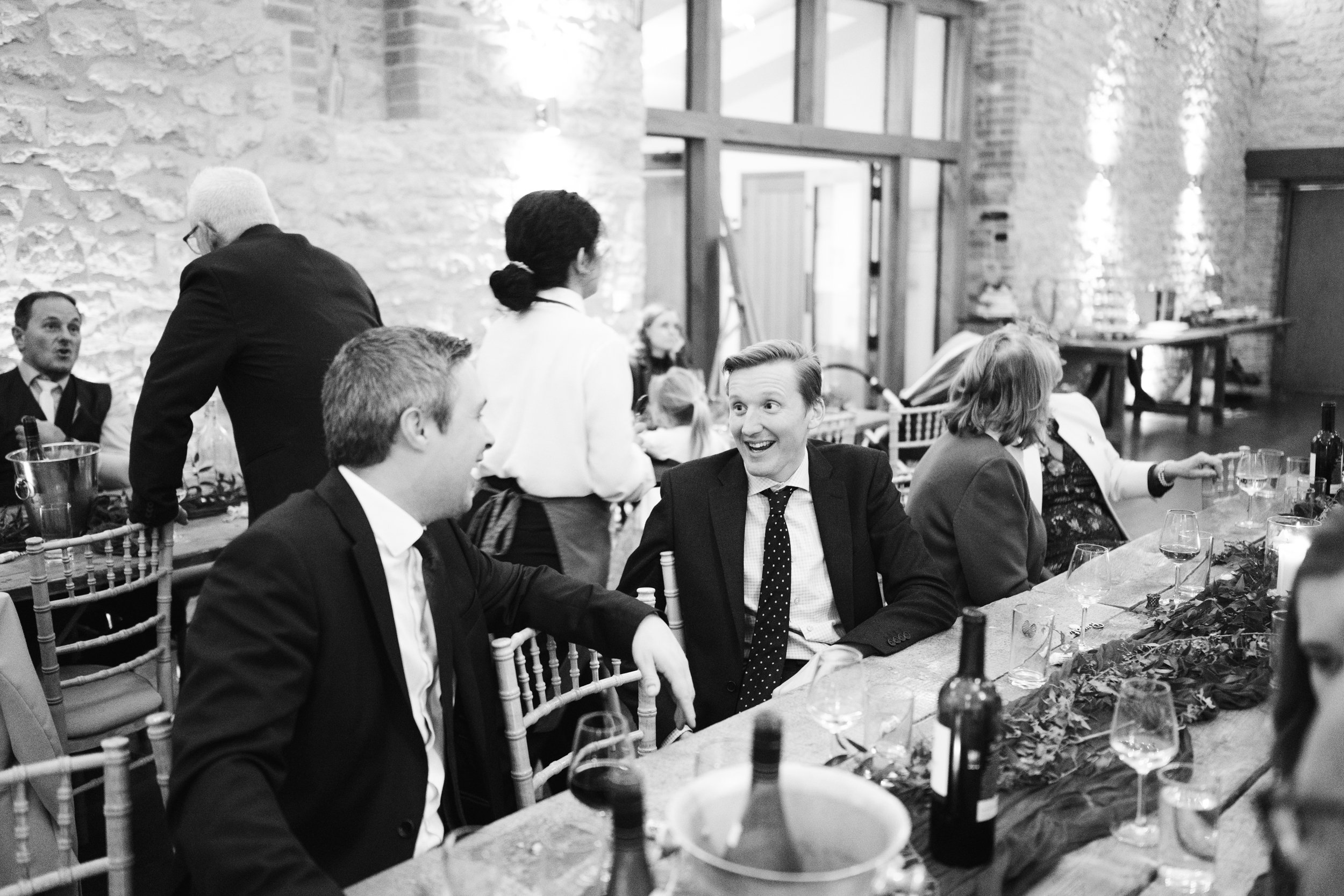 People sitting at a long dining table with wine glasses, bottles, and decorations during a social gathering or celebration in a rustic venue with brick walls and large windows.