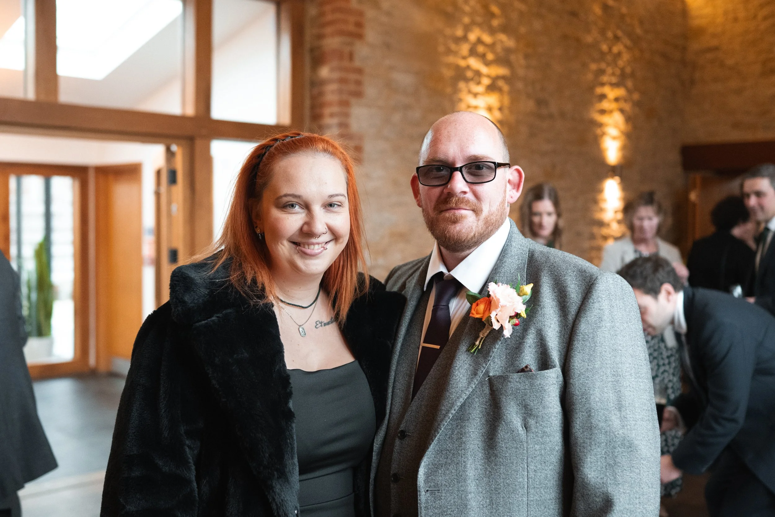 A smiling woman with red hair and a man with glasses and a beard dressed in a grey suit with a boutonniere at a social event.