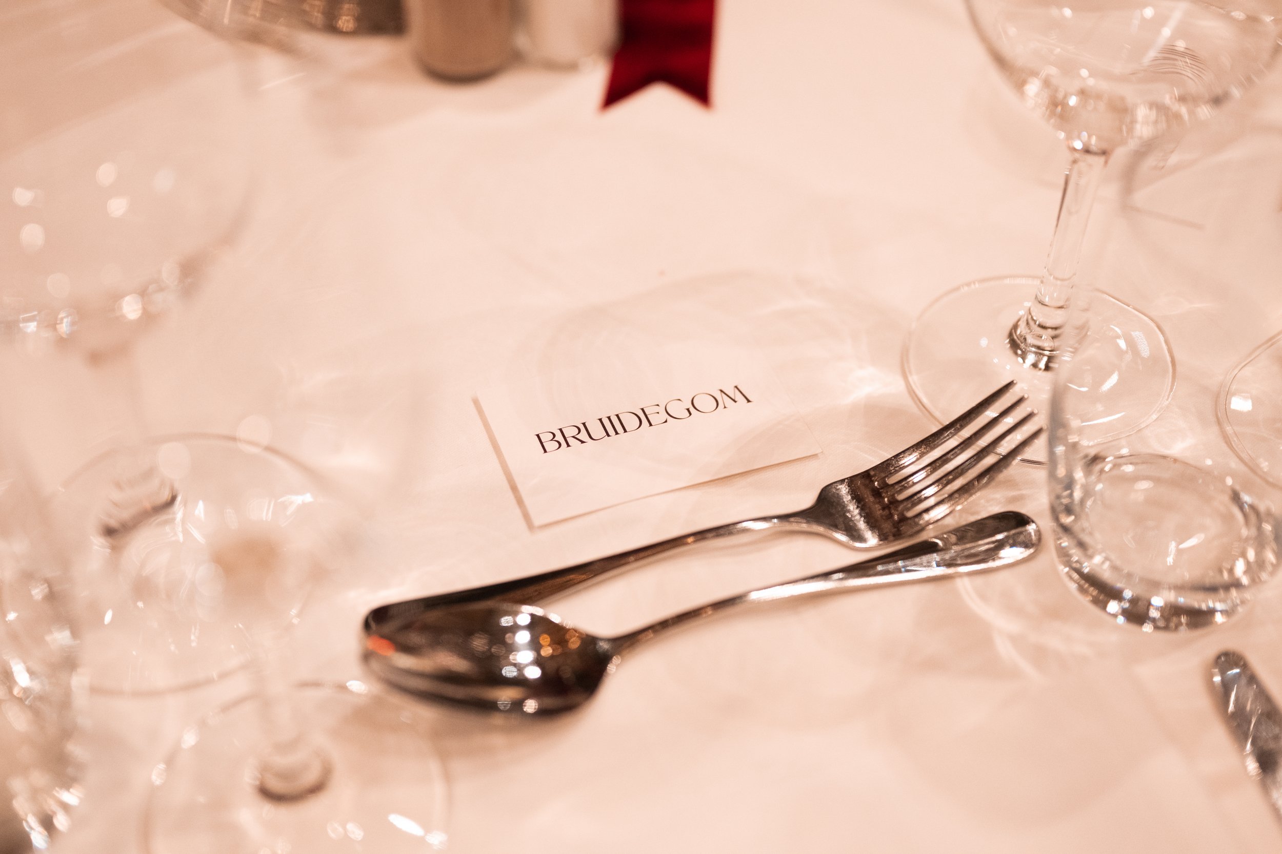 Table setting with a place card labeled 'BRUIDEGOM,' forks, spoons, and empty wine glasses on a white tablecloth.