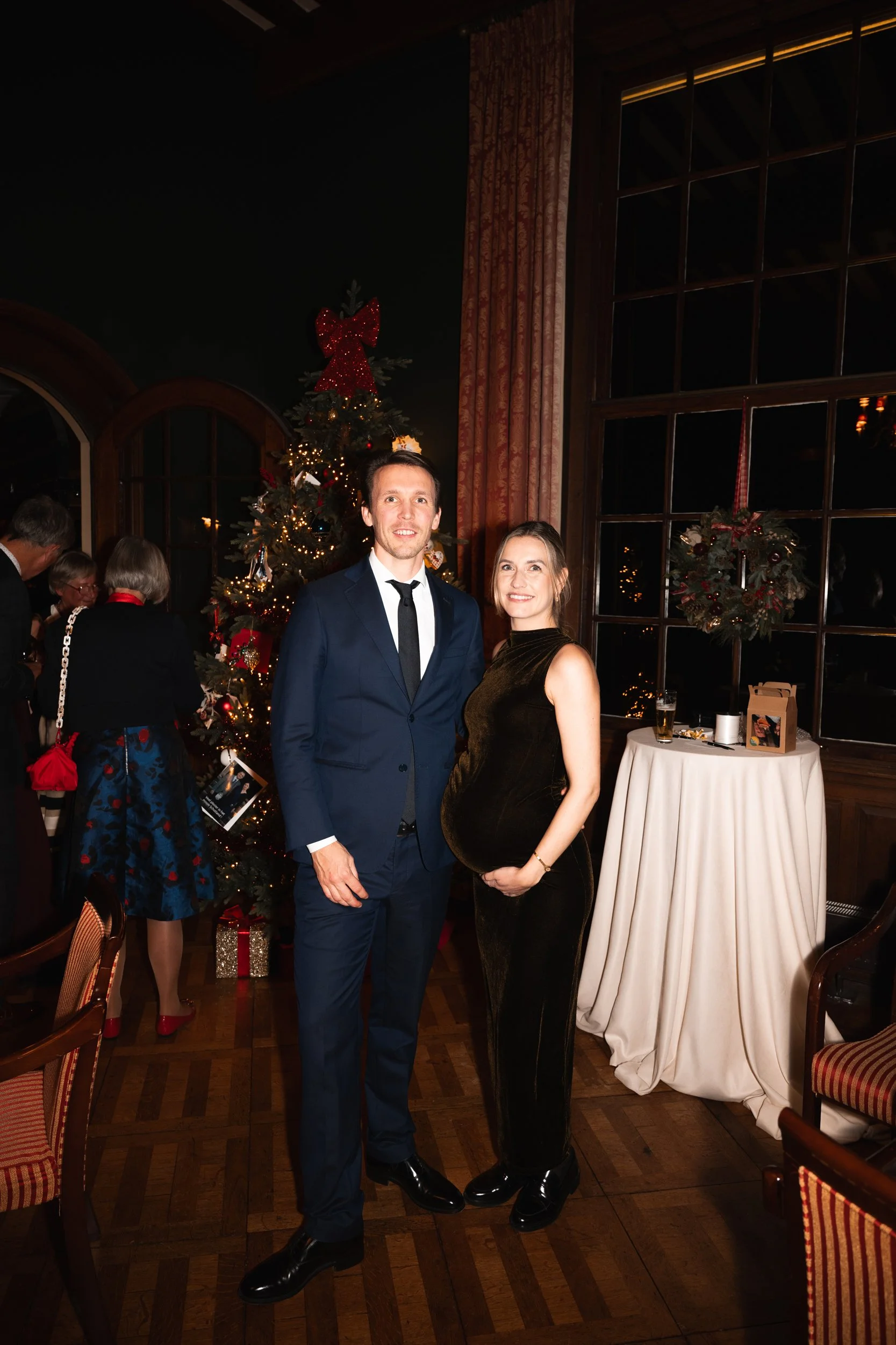 A man in a blue suit and a woman in a black dress are posing together during a holiday party in front of a decorated Christmas tree, with other guests in the background.