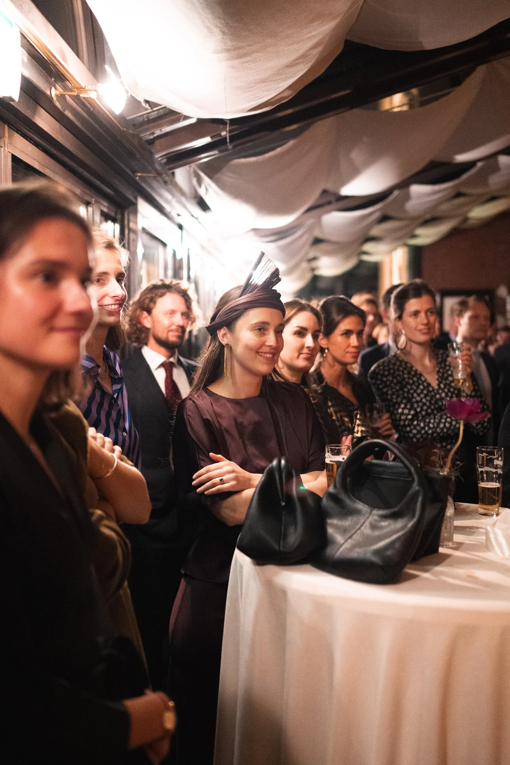 Group of people attending a formal event, standing around a round table decorated with a flower and black bags, in an indoor venue with draped ceiling fabric.