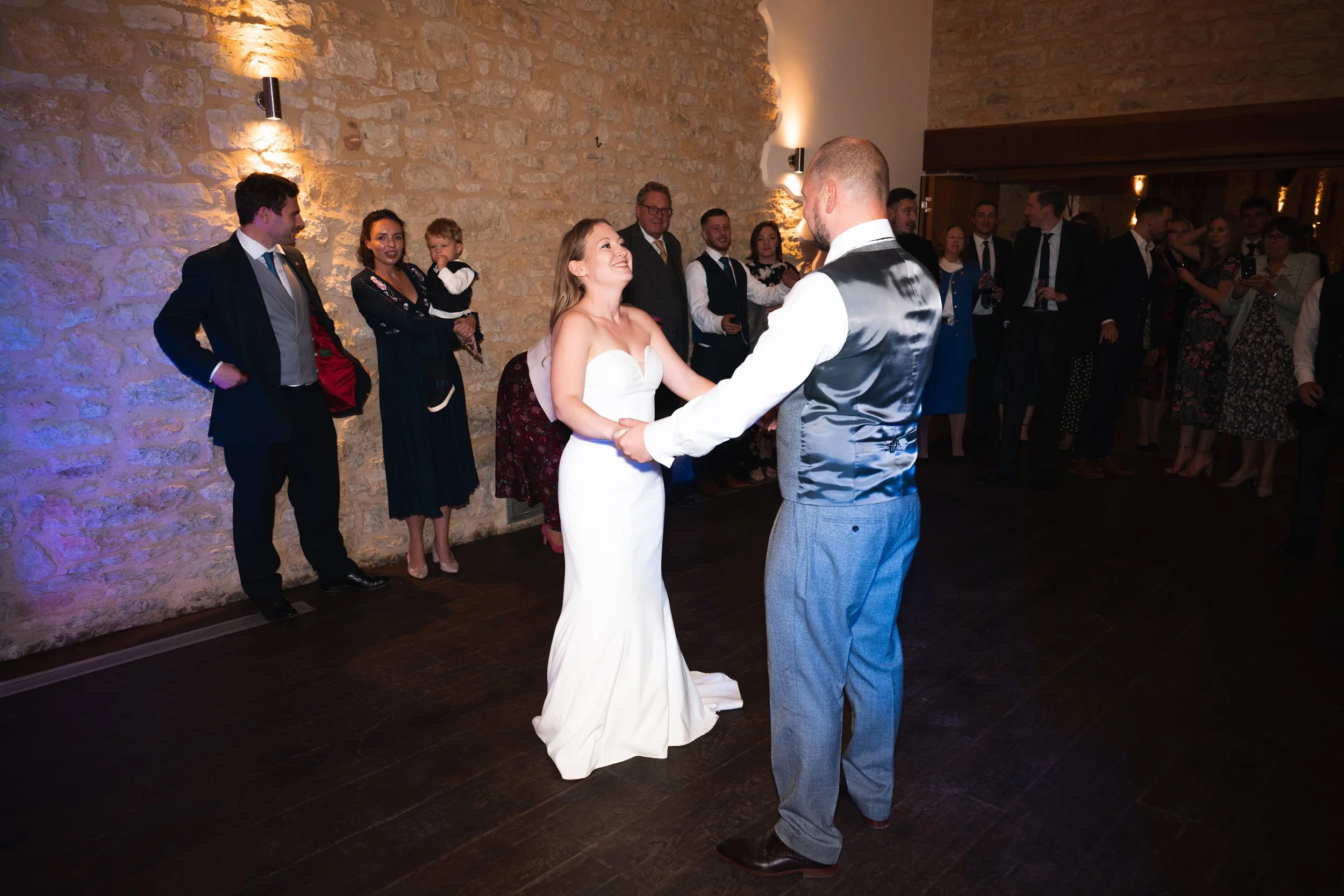 A bride and groom are dancing and holding hands while smiling at each other during their wedding reception. There are guests in the background watching the couple dance, some with drinks in hand, standing against a stone wall with warm lighting.