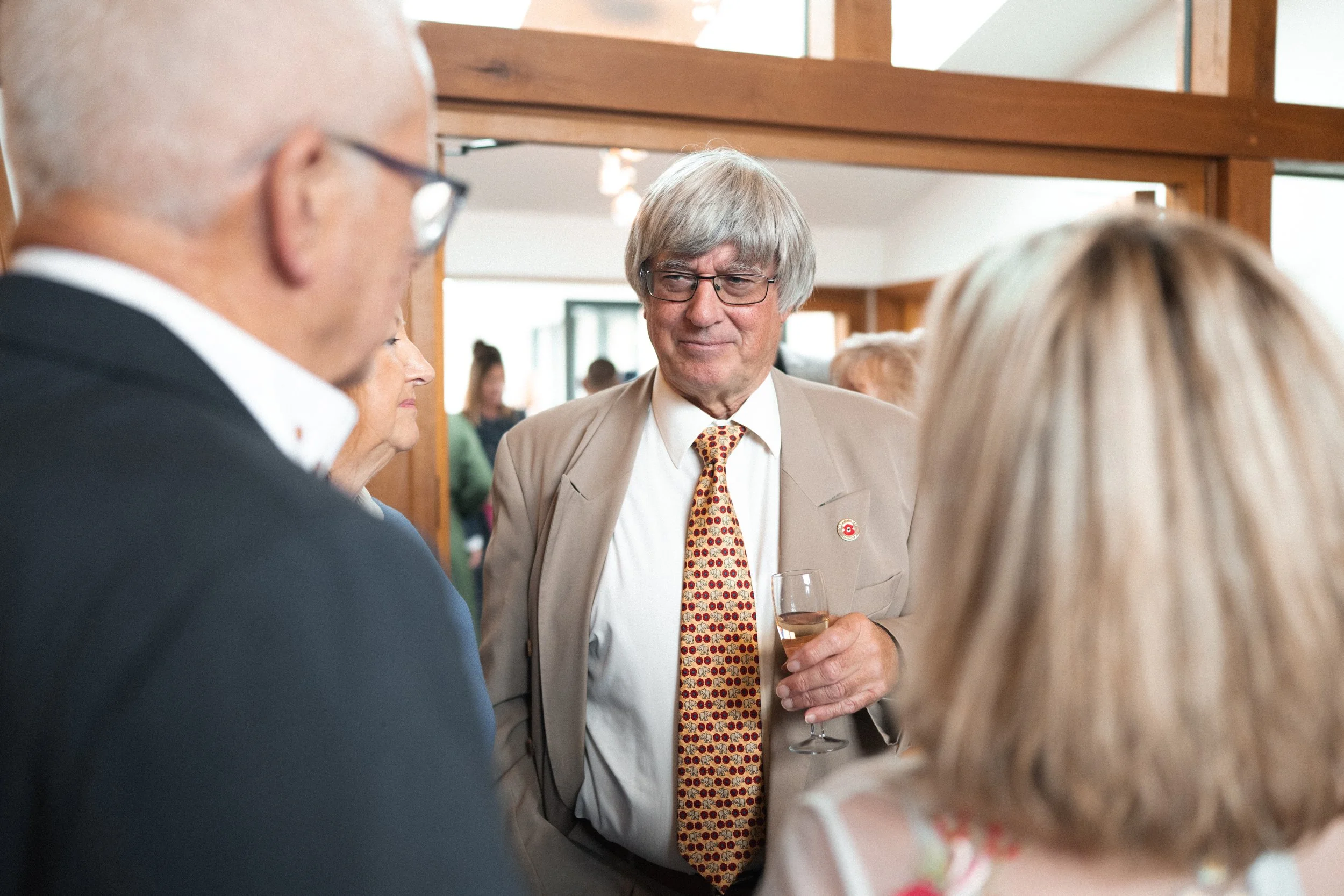 A group of people in conversation at a social gathering, with a man holding a glass of wine in the foreground, blurry figures in the background, and wooden decor.