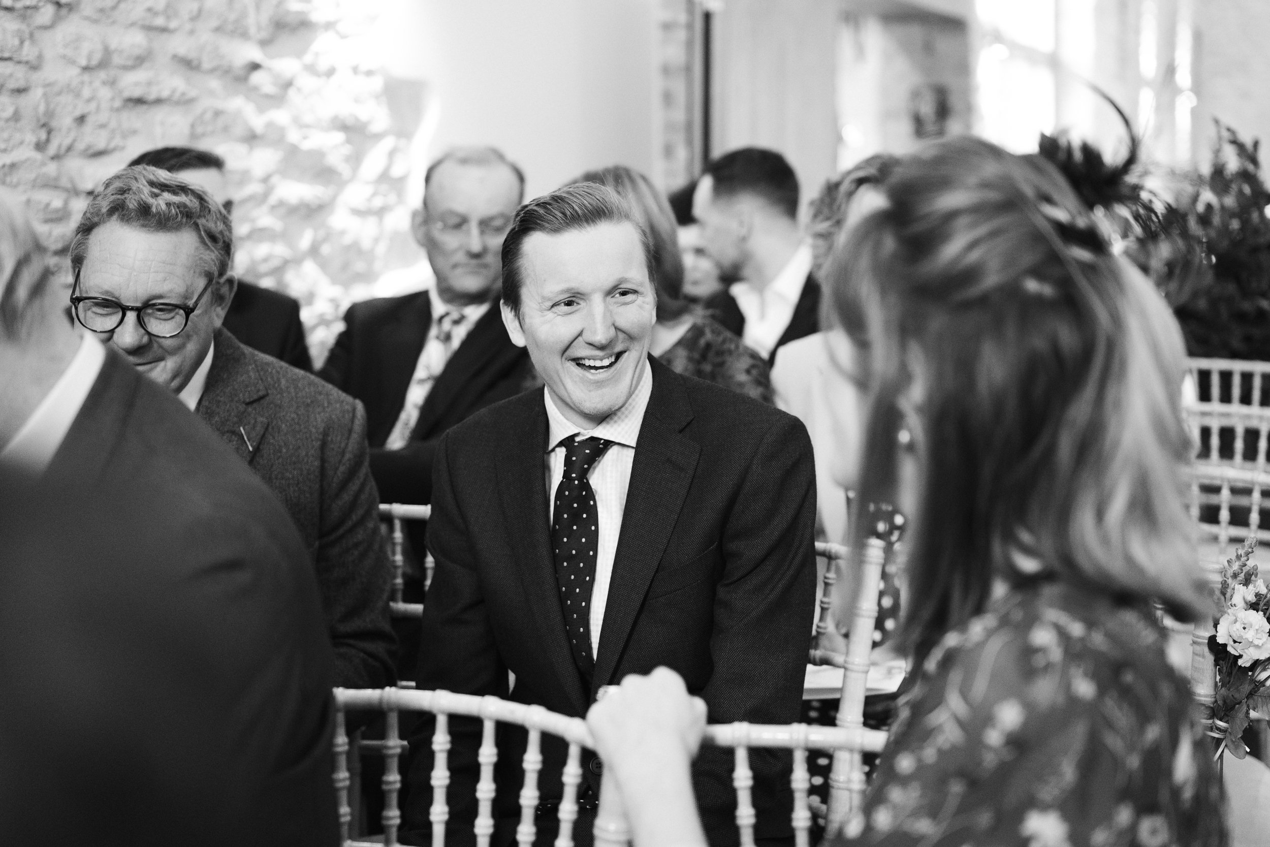 Black and white photo of a group of people at a social event, with a man in a suit and polka dot tie smiling and engaging in conversation, surrounded by other well-dressed individuals seated and interacting.