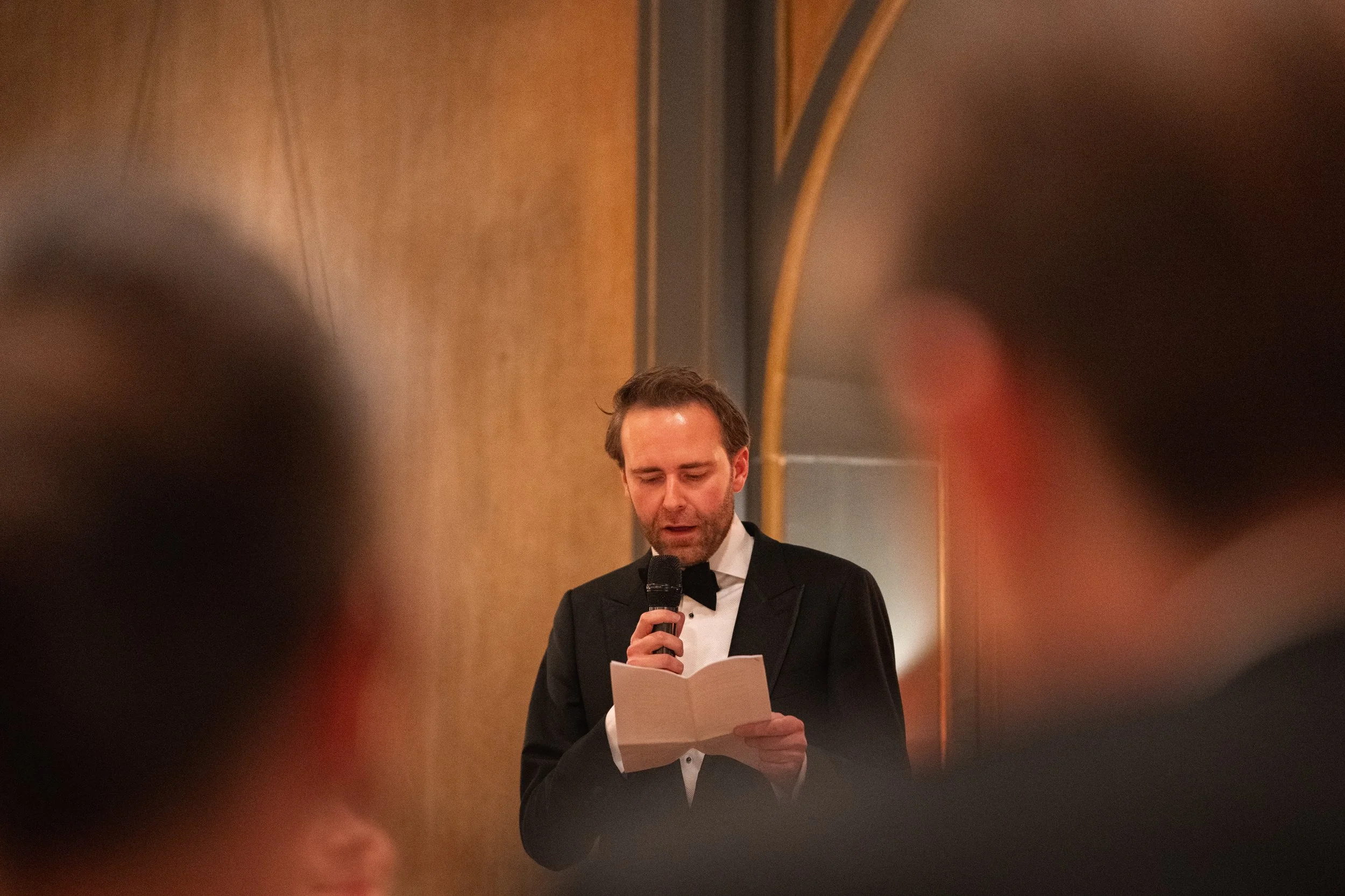 A man in a tuxedo with a bow tie is reading from a piece of paper while speaking into a microphone at a formal event.