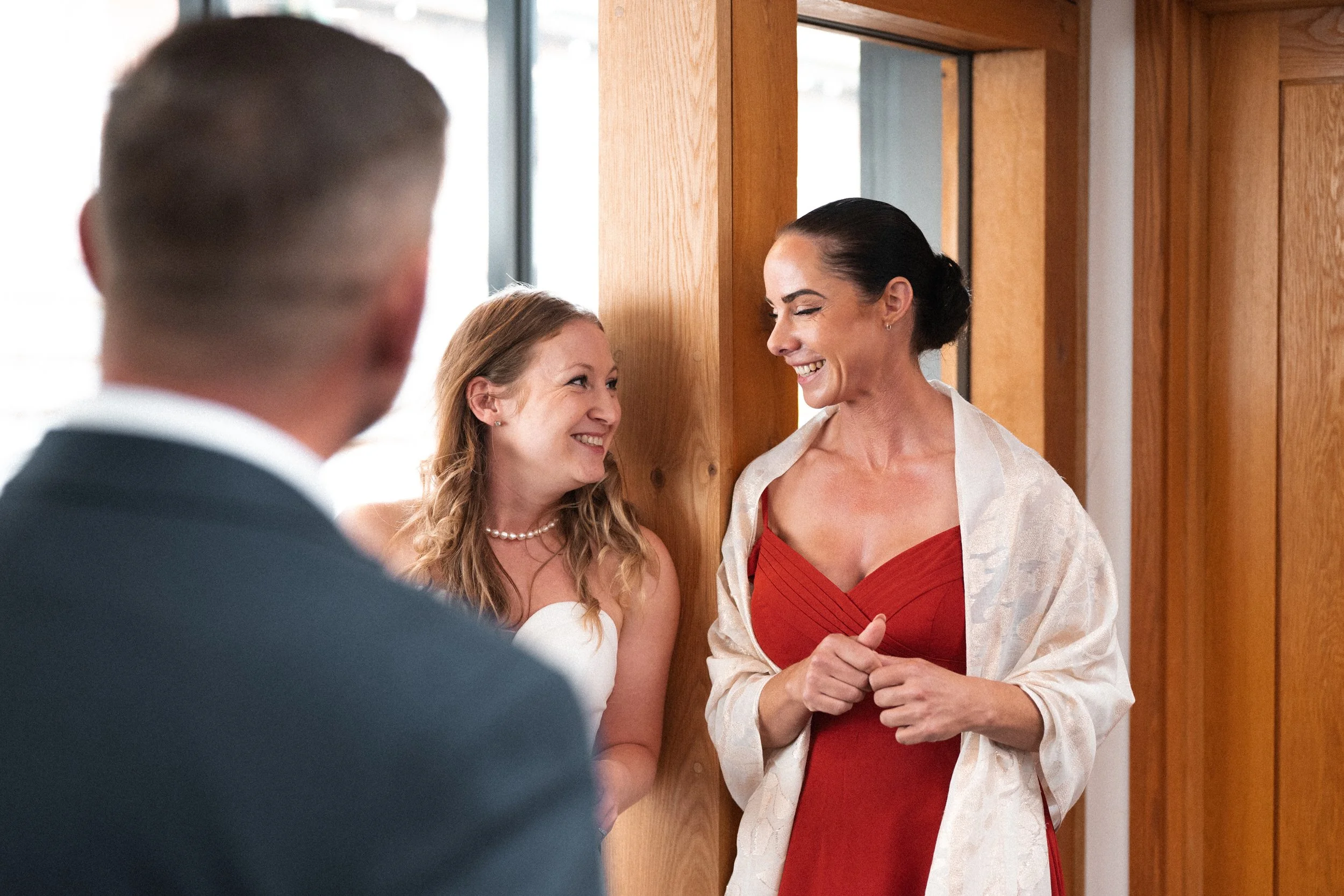 Two women smiling and talking near a doorway, with a man in a suit in the foreground. One woman is wearing a white dress with a pearl necklace, and the other woman is in a red dress with a cream-colored shawl.