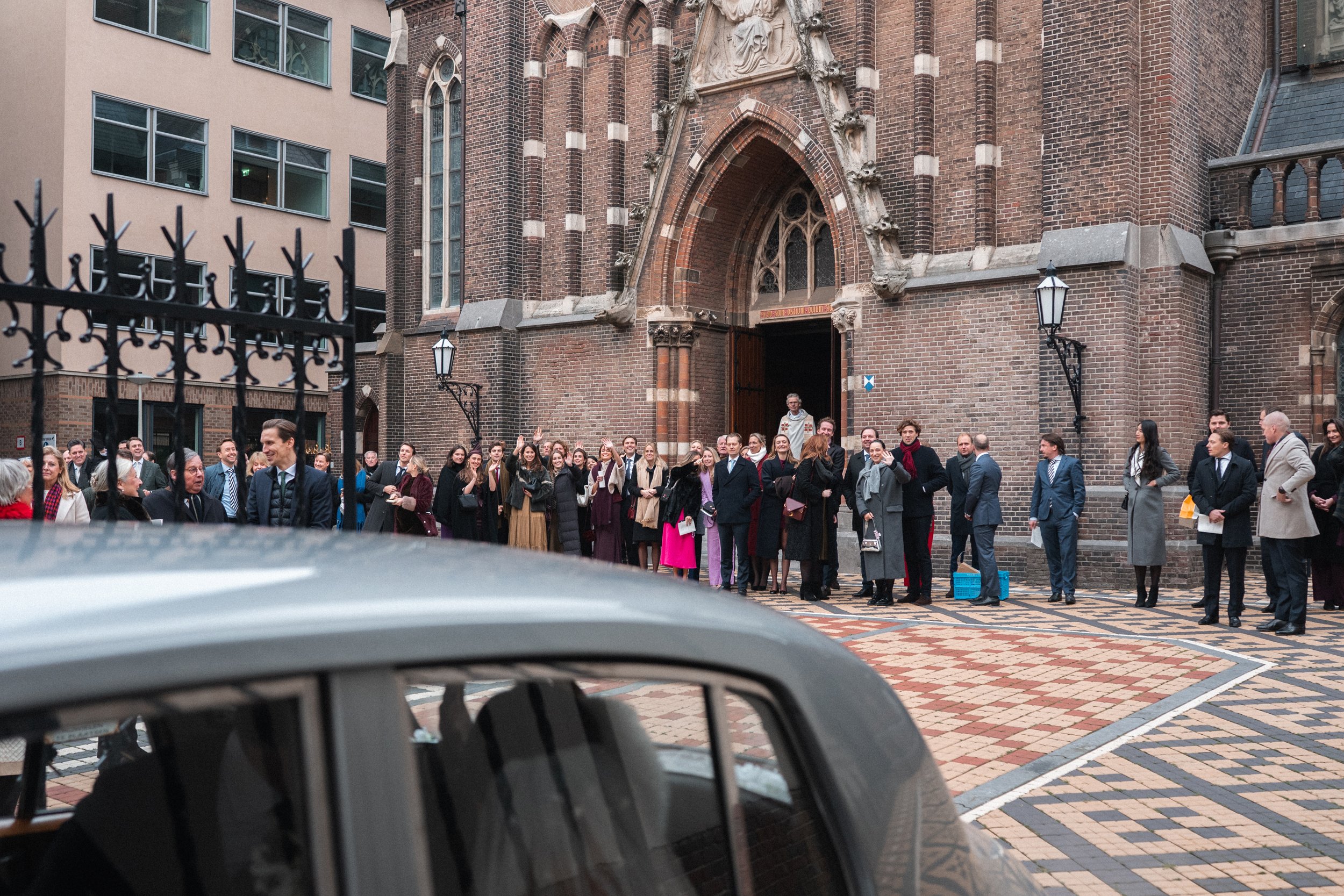 People standing outside a large brick church with an arched entrance, waiting in a line, dressed in formal attire, with a vintage car in the foreground.