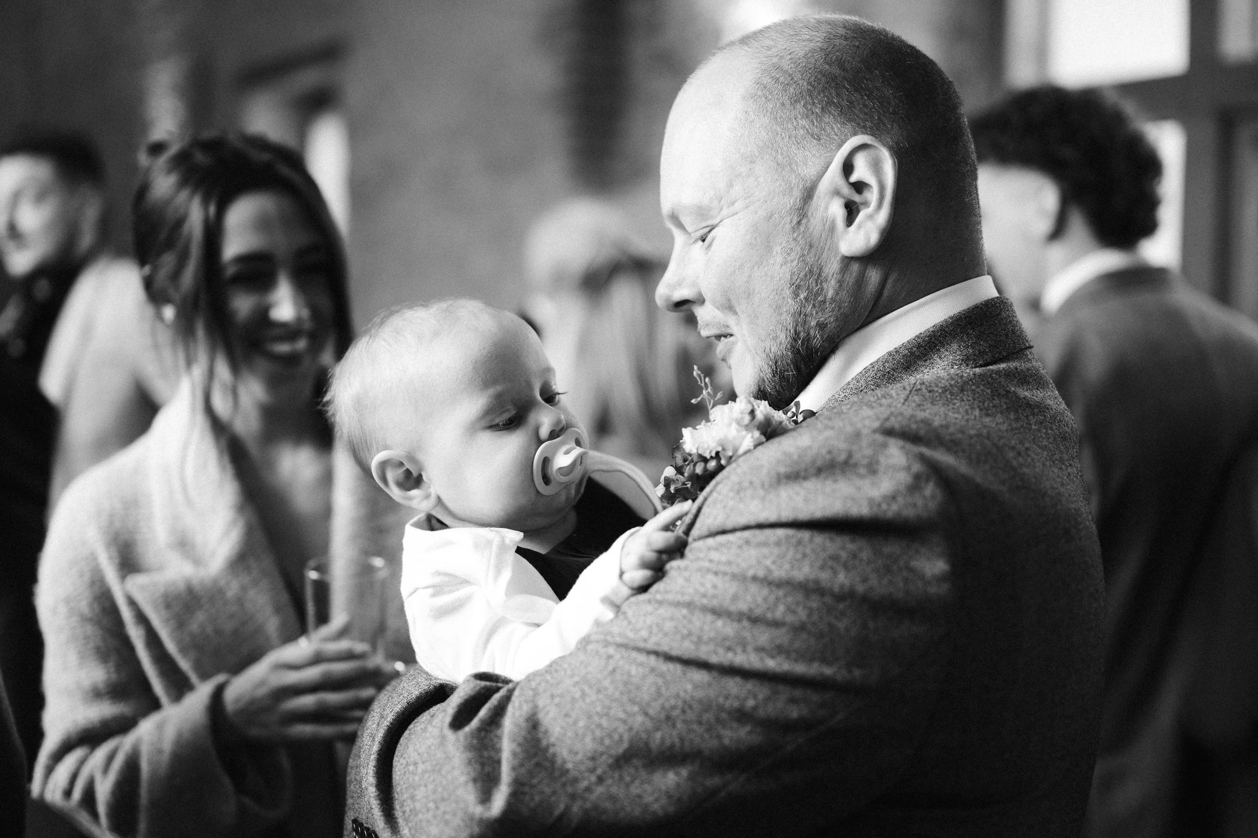 A man in a suit holding a young child with a pacifier, smiling as they look at each other; in the background, a woman holding a glass, smiling, with other people at what appears to be a social gathering.