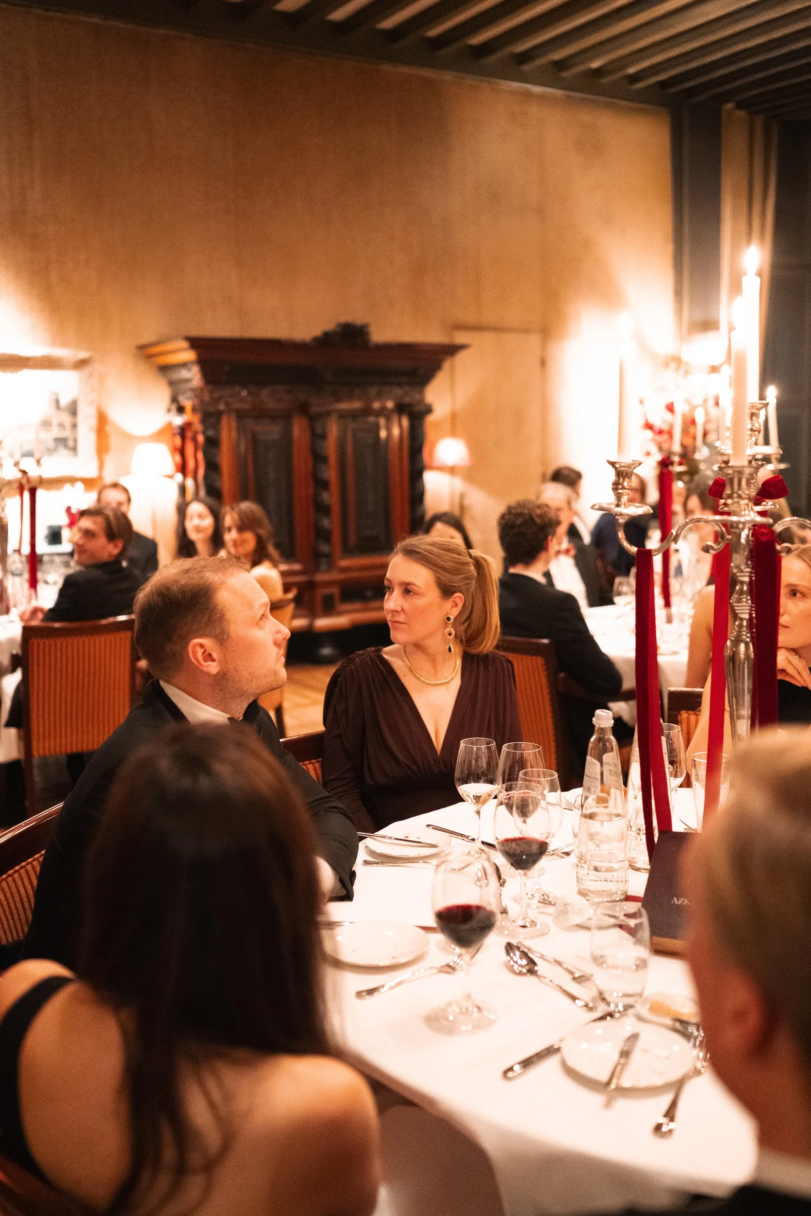 Guests seated at a dinner table with wine glasses, water bottles, and silverware during a formal event in an elegant room decorated with candles and red ribbons.