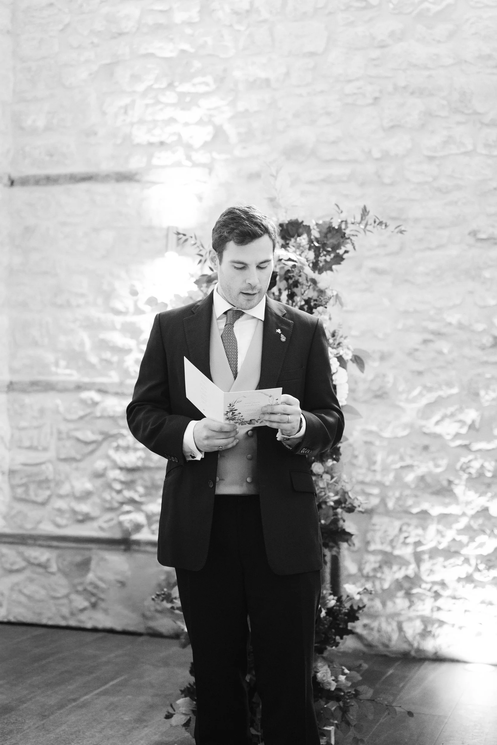 A man in a suit and tie reading a card, standing in front of a floral arrangement and a stone wall.