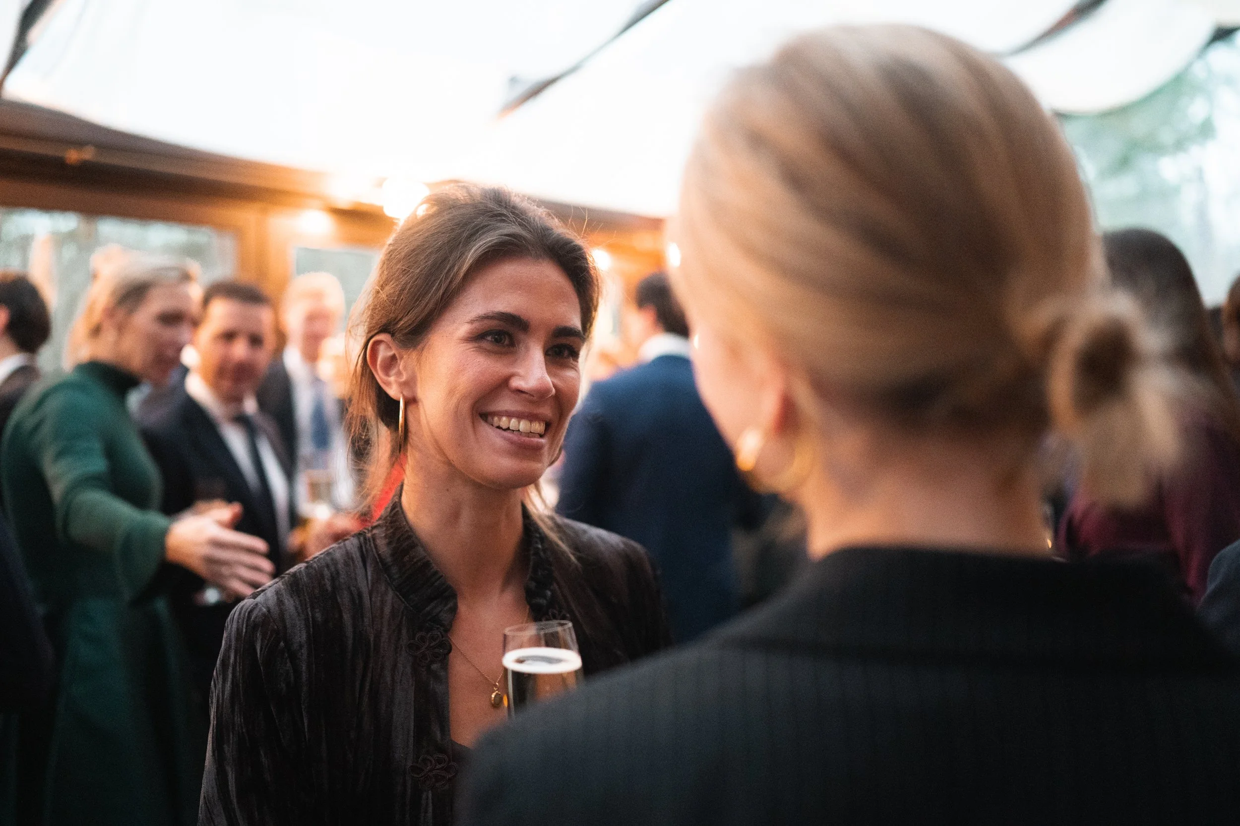 Two women conversing at a social event, with one smiling and holding a glass of champagne, while other guests are mingling in the background.