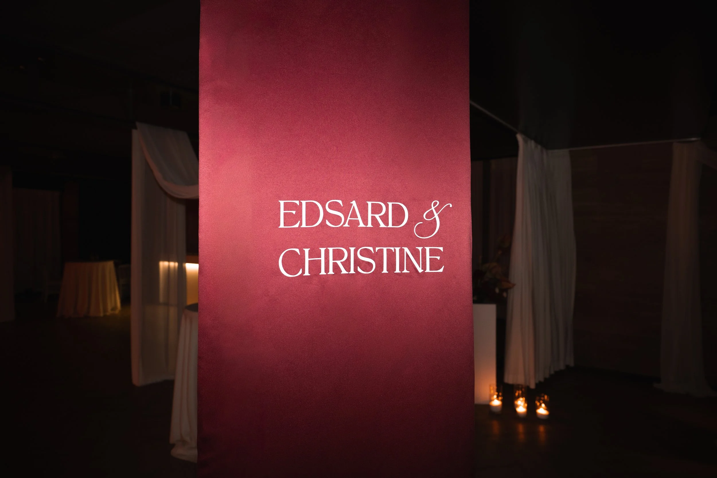 A pink vertical banner with white text reading 'Edsard & Christine' at an indoor event with dim lighting and candles in the background.