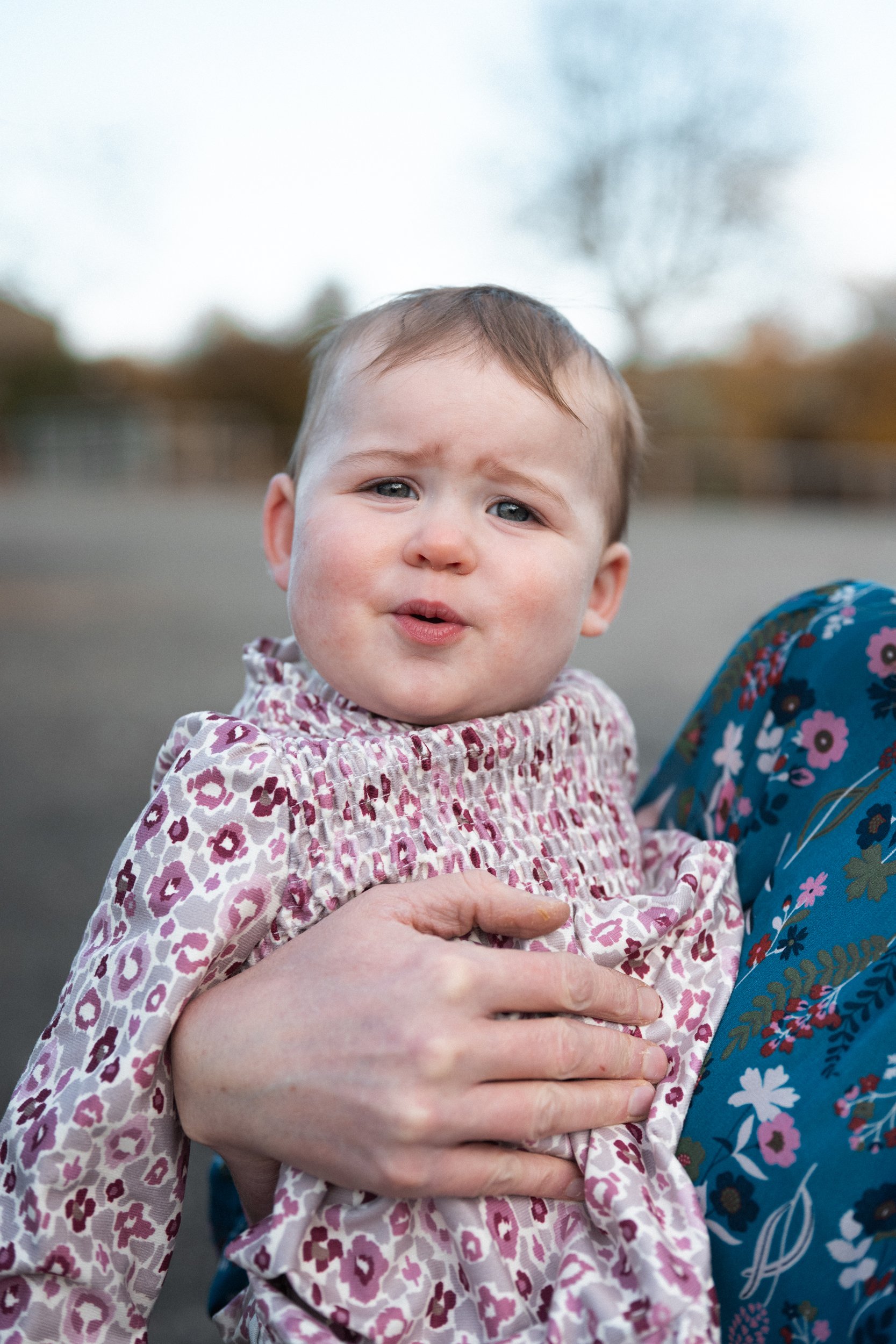 A young child with a distressed facial expression being held by an adult. The child is wearing a pink and white patterned top, and the adult's hand is visible, holding the child's arm. The background appears to be an outdoor setting with blurred tree
