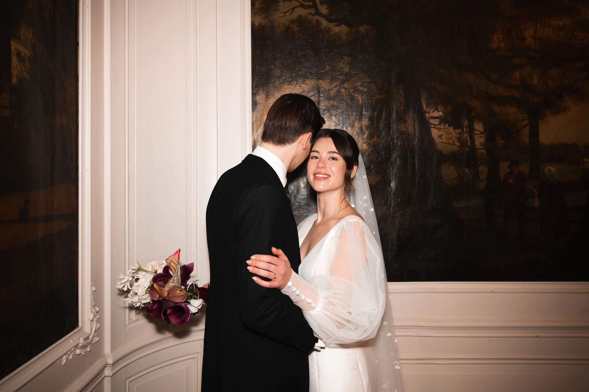 A bride and groom celebrating marriage indoors, with the bride smiling and holding a bouquet of flowers, standing next to a large dark painting or window.