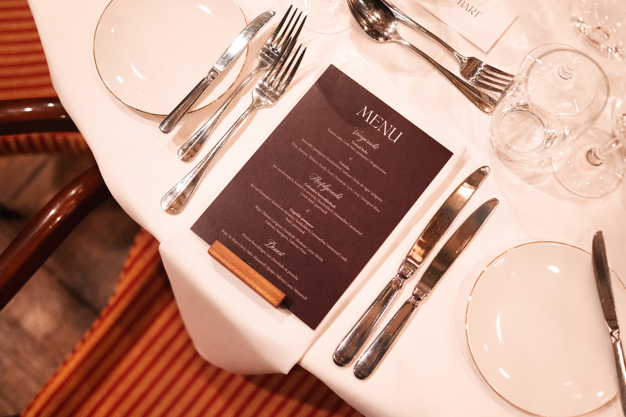A formal dining table with a black menu card, silverware including knives and forks, empty white plates with gold rims, and upside-down wine glasses, set on a white tablecloth.