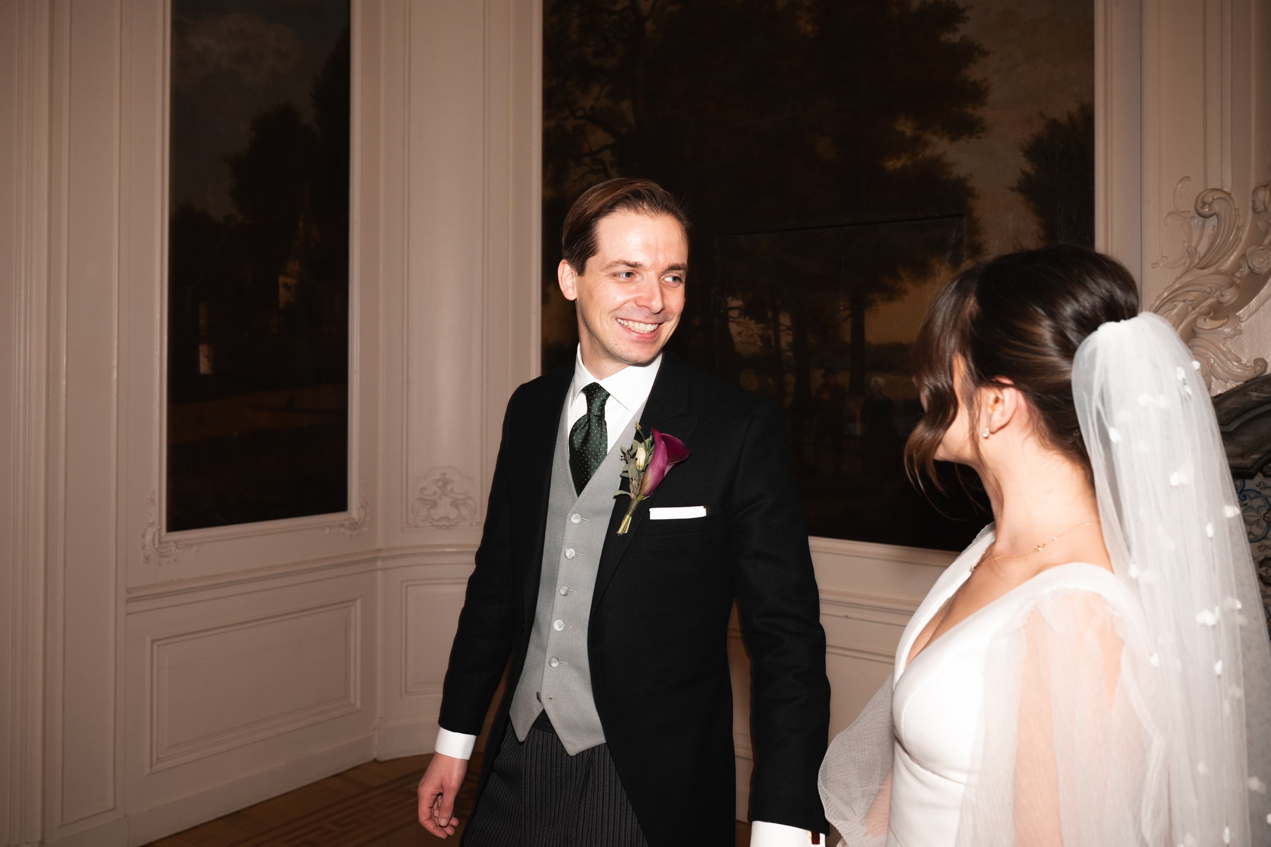 A smiling groom in a black tuxedo with a boutonniere, standing and looking at a bride with a veil, in a decorated elegant room with large framed artwork.