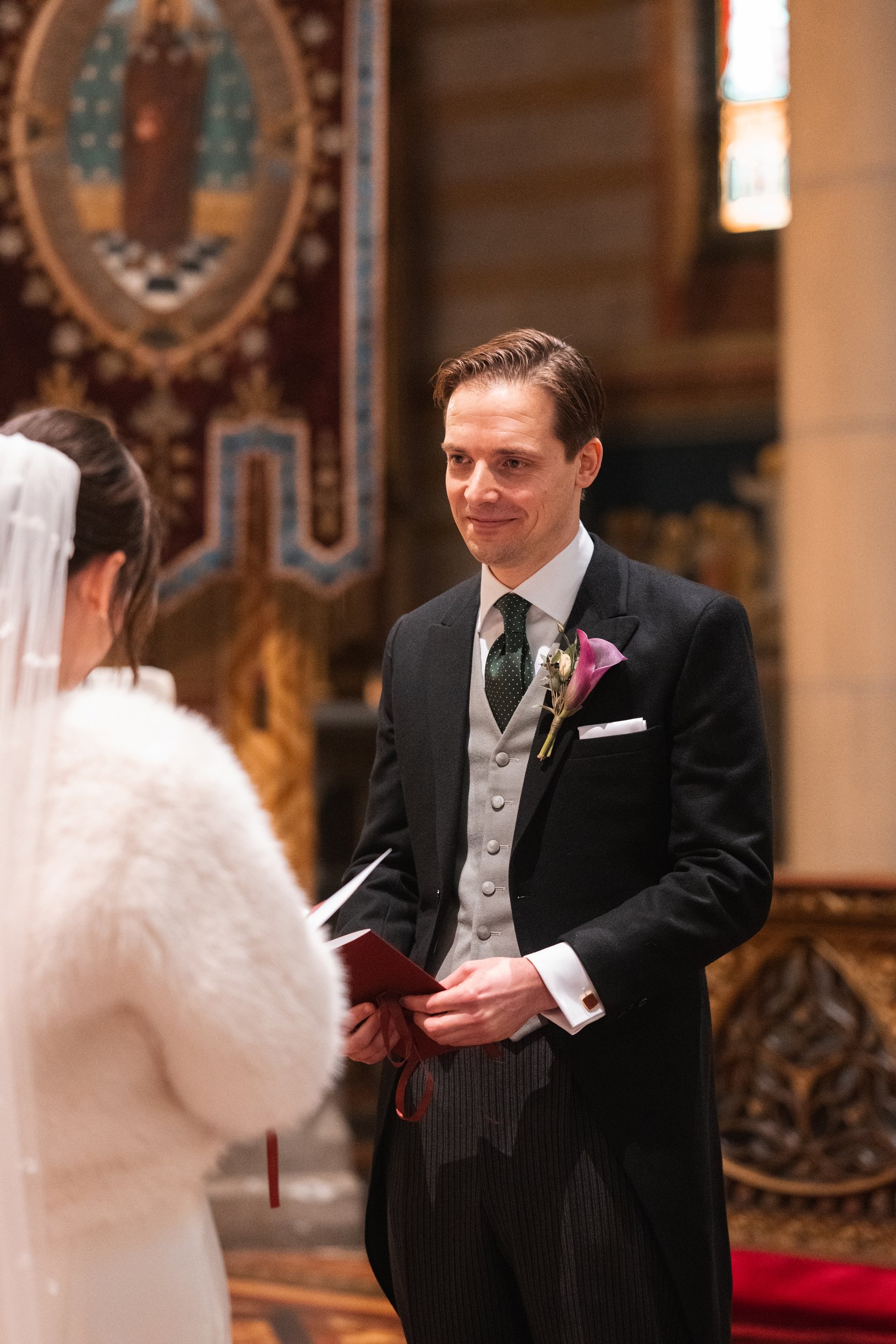 A groom dressed in a black tuxedo with a boutonniere, standing before a bride in a white dress and veil during a wedding ceremony inside a church with religious artwork and wooden decor.