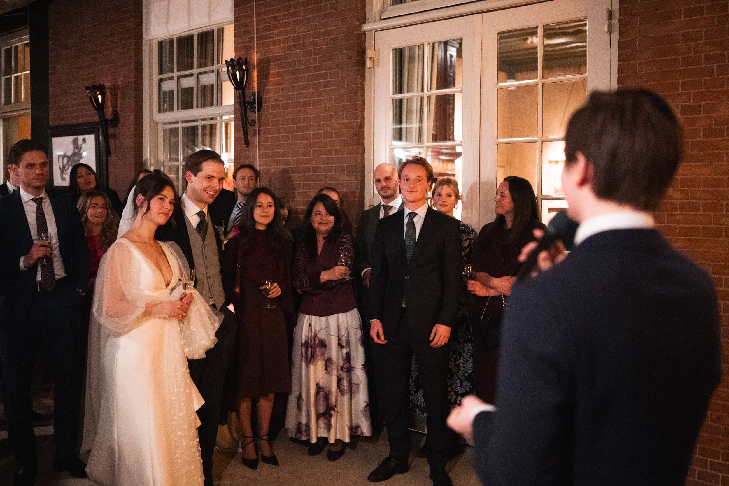 People attending a wedding reception, listening to a speech inside a brick-walled venue with large windows and warm lighting.