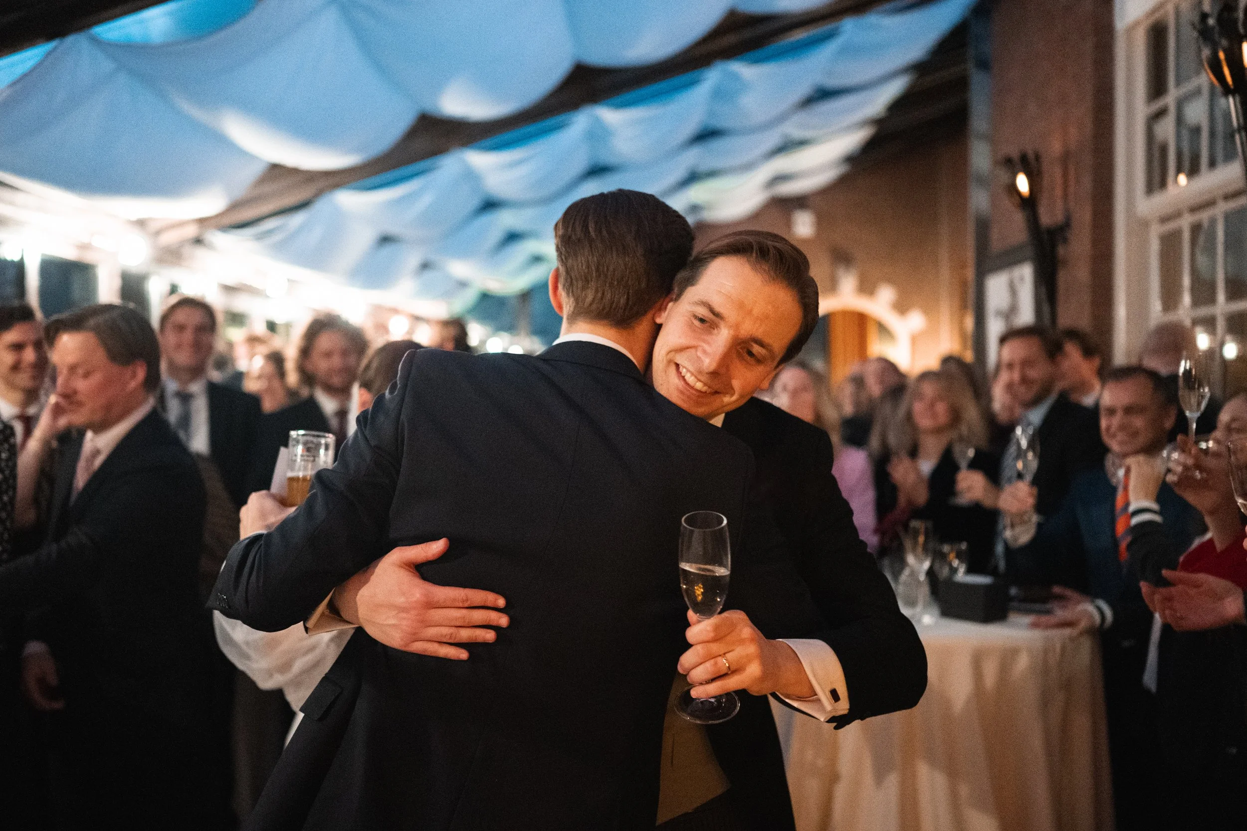 Two men hugging at a celebration or party, one holding a glass of champagne, with a crowd of people in the background.