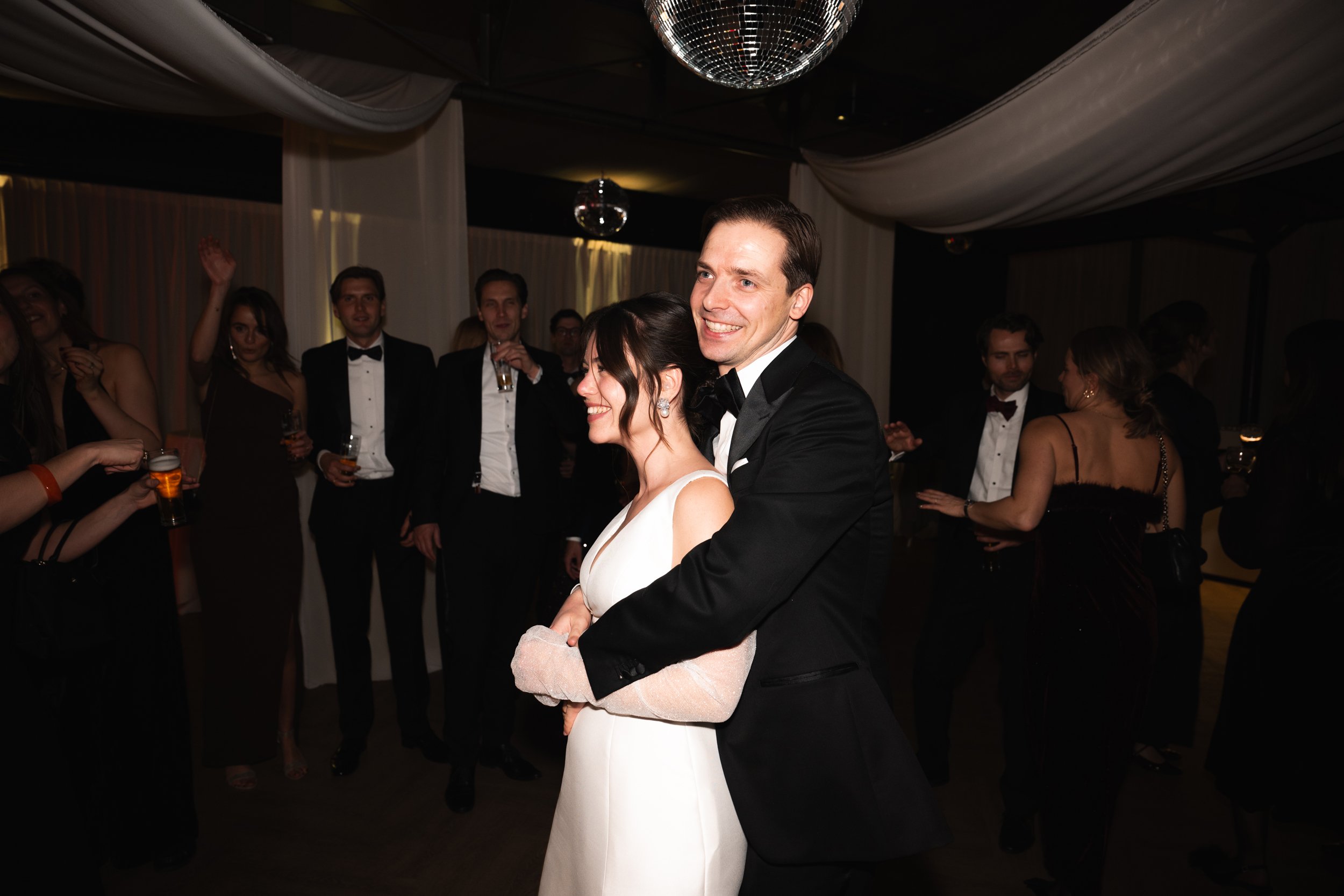 A bride and groom dancing at their wedding reception, surrounded by guests, with a disco ball overhead and elegant drapery decorations.