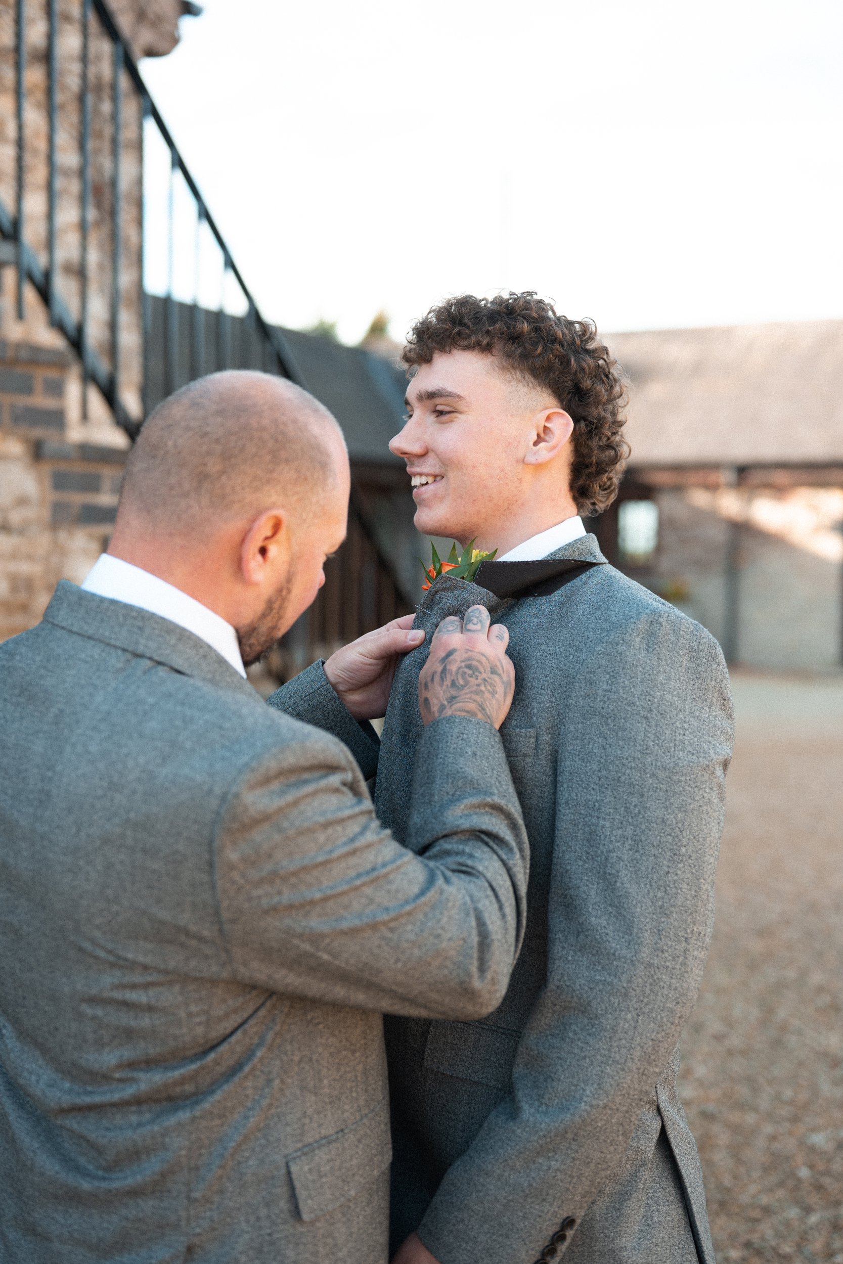 A man helping another man adjust a boutonniere on his tuxedo jacket during a wedding, outdoors near brick buildings.