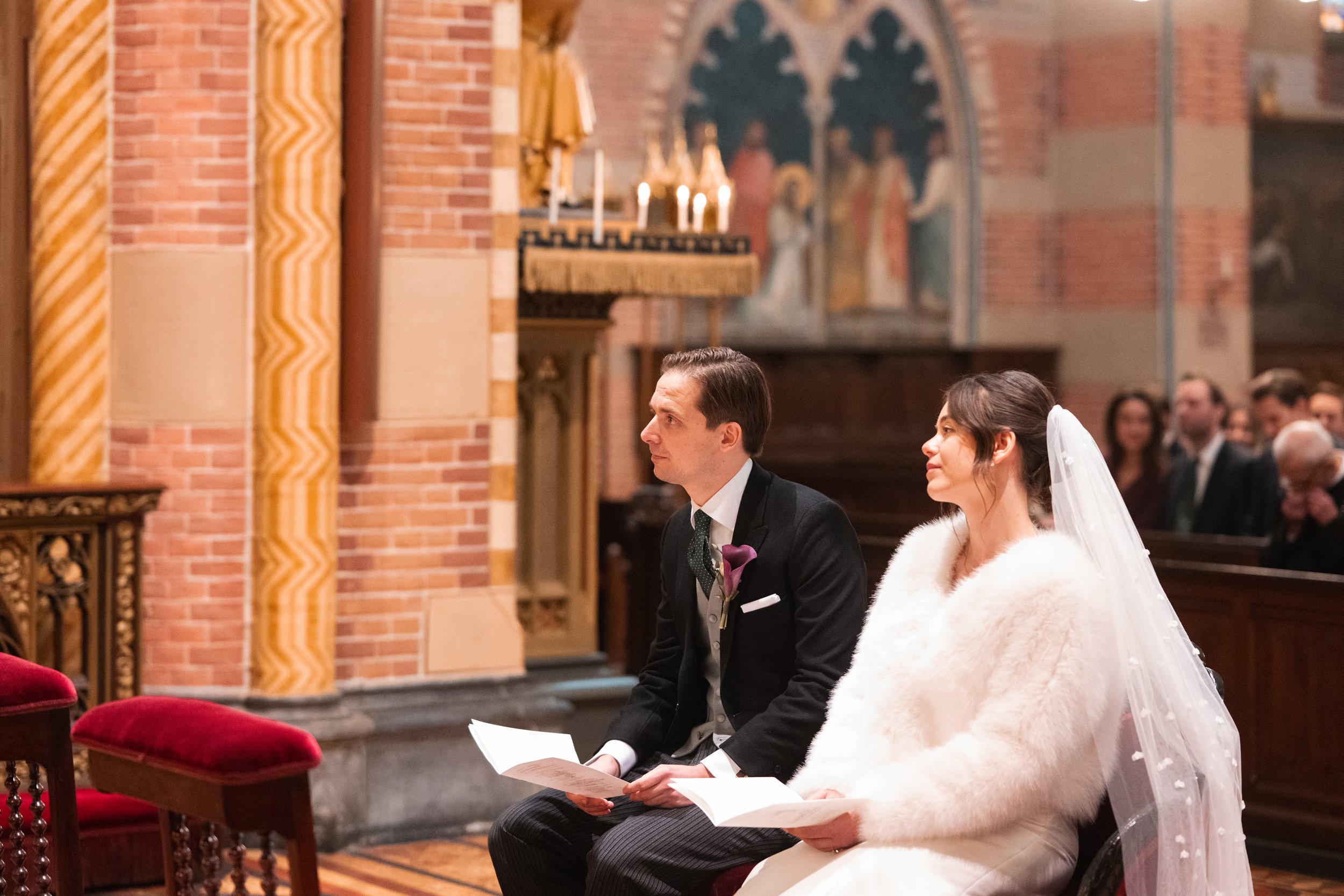 A bride and groom seated in a church during their wedding ceremony, the bride wearing a veil and white fur coat, and the groom in a black tuxedo, with guests visible in the background.