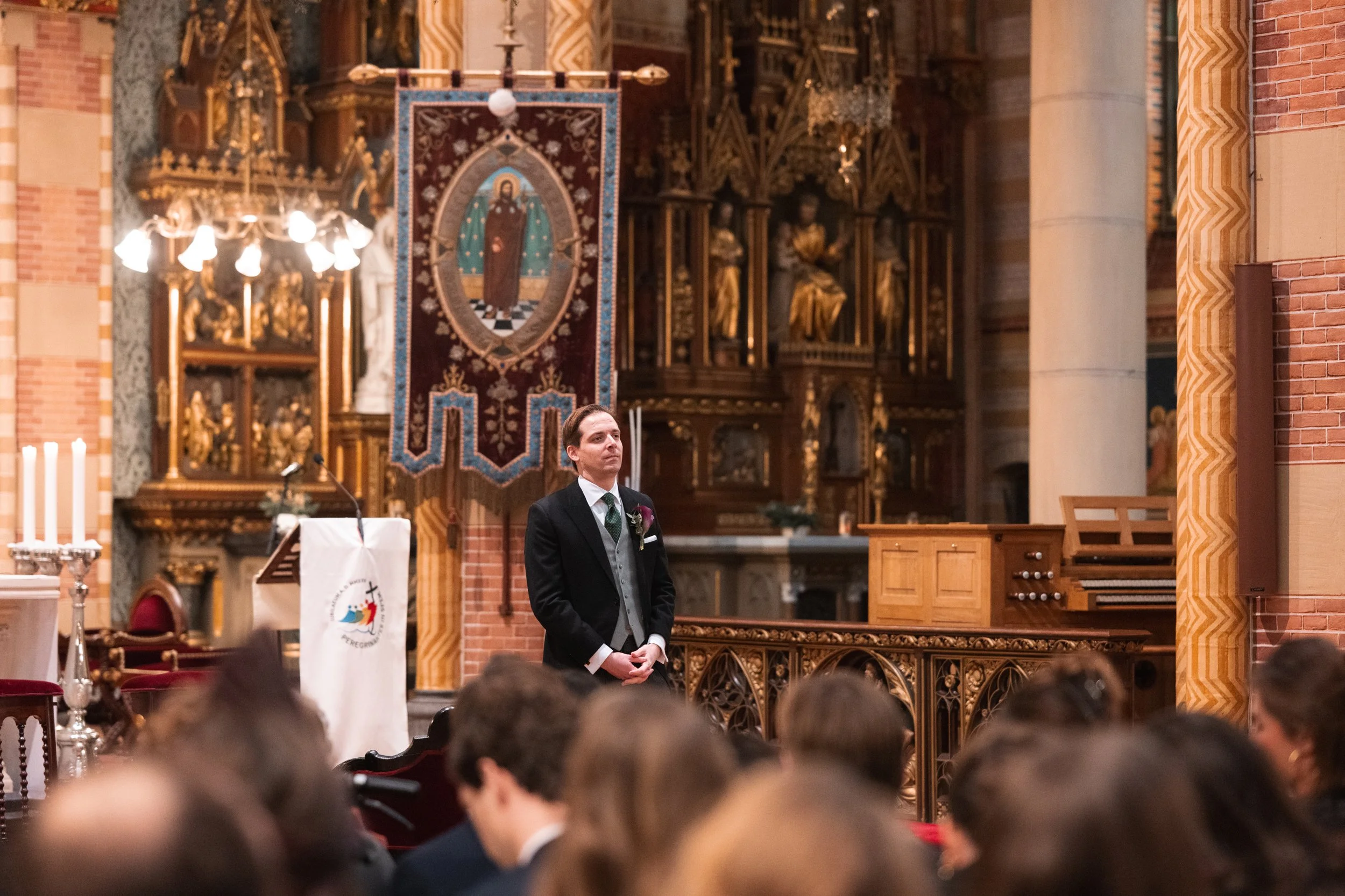 A man in formal attire standing in a church during a wedding ceremony, with guests seated in front of him and ornate religious decorations and banners behind.