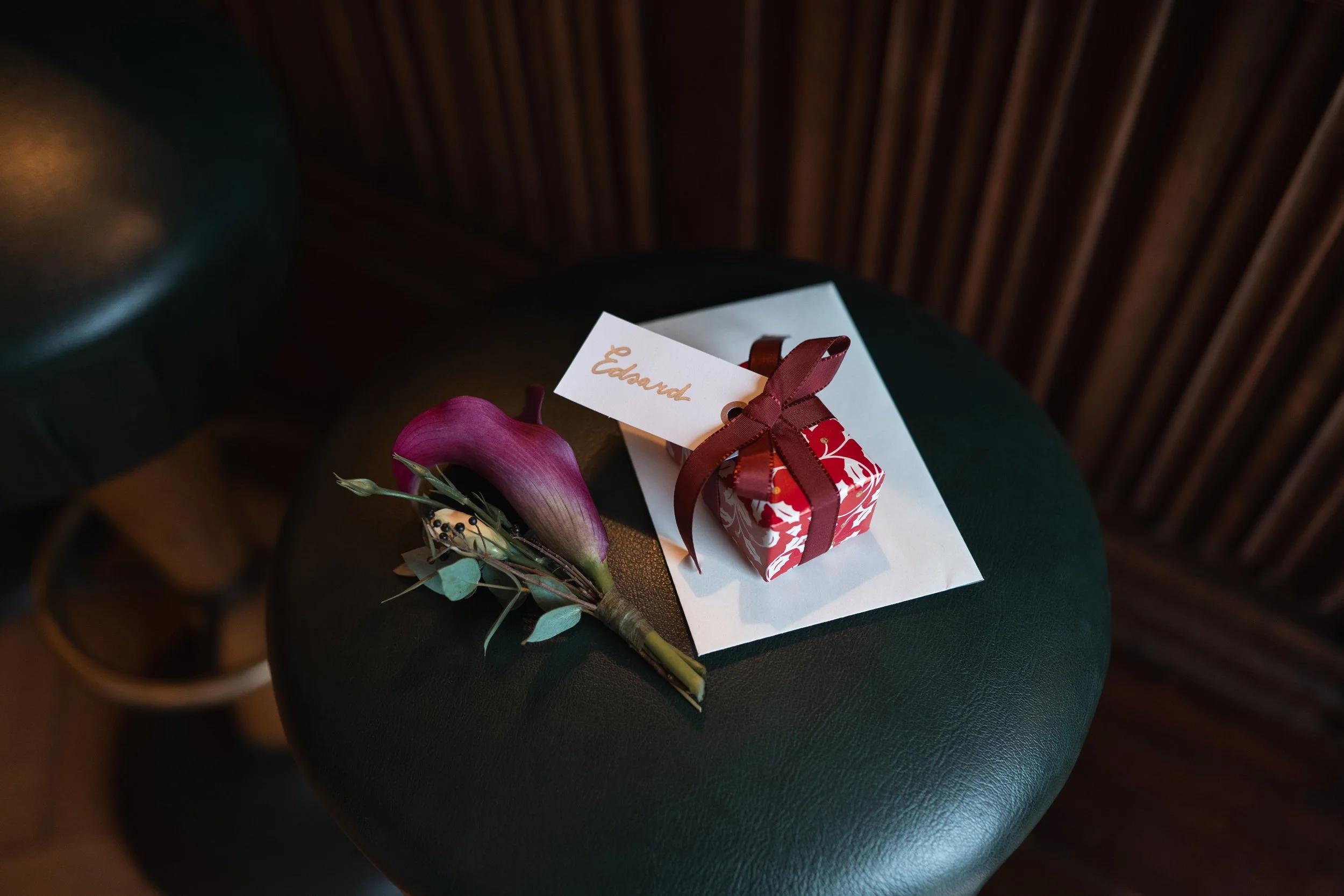 A small, red and white floral patterned gift box with a brown ribbon and a gift tag that says "Eward" is on a white paper sheet. Next to it, there is a single pink calla lily flower with green leaves and dried foliage, all placed on a dark green roun