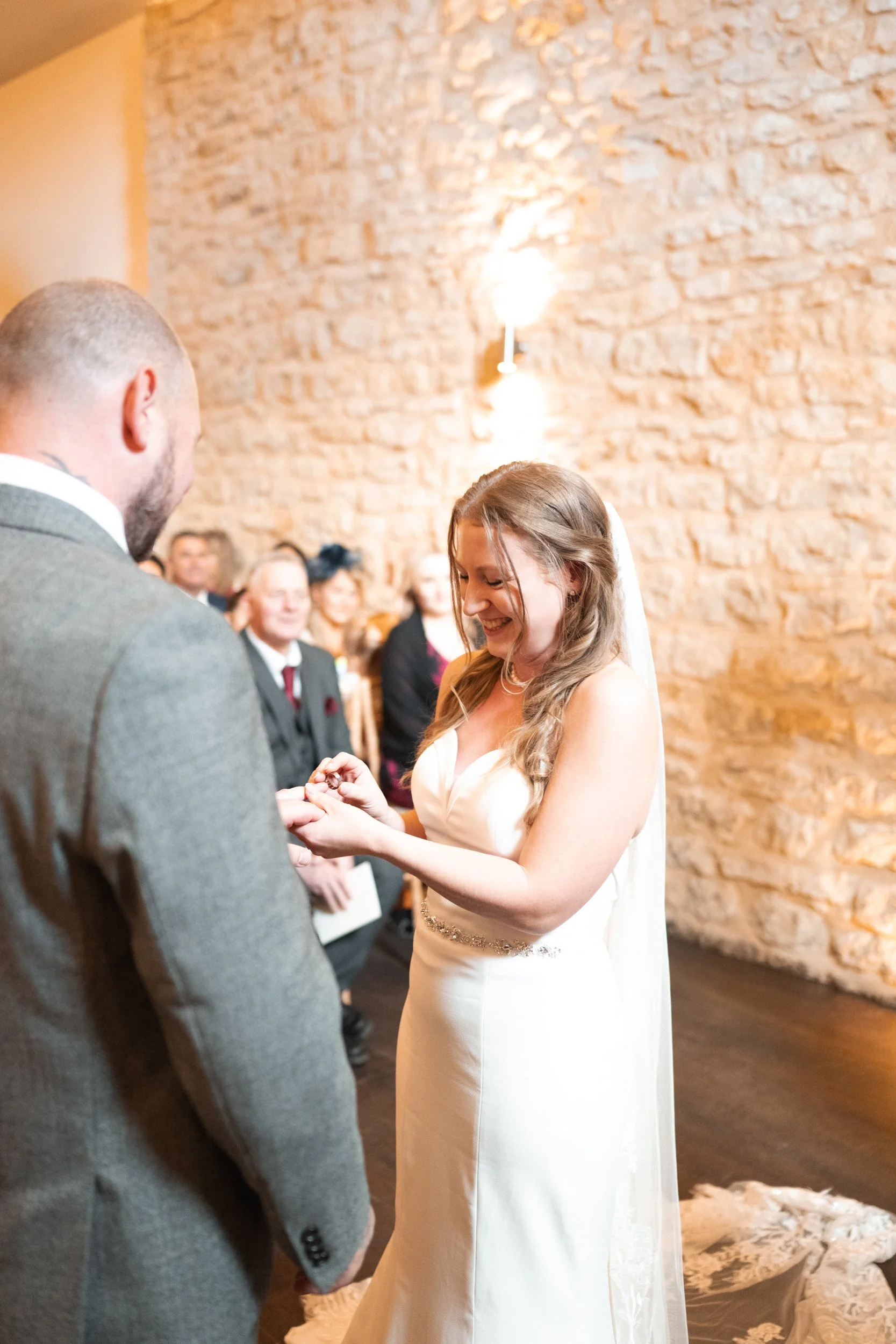 Bride and groom exchanging rings during their wedding ceremony in a rustic indoor setting with an exposed brick wall and seated guests in the background.
