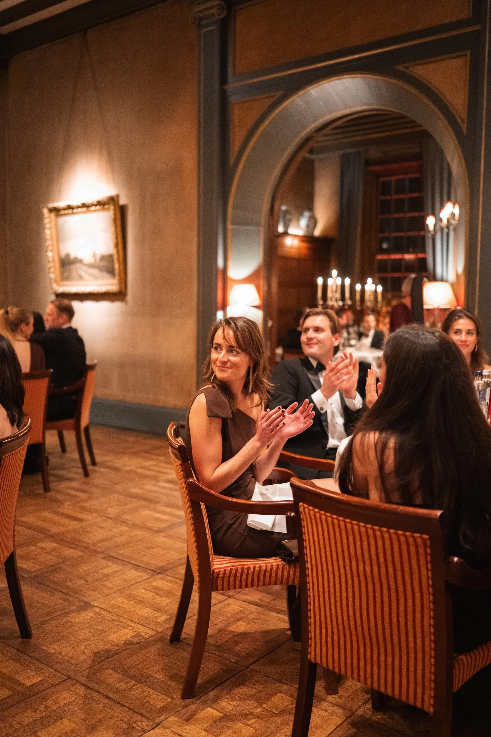 People dressed in formal attire sitting at a dining table, clapping and smiling during an event in an elegant, warmly lit room with artwork and candelabra.