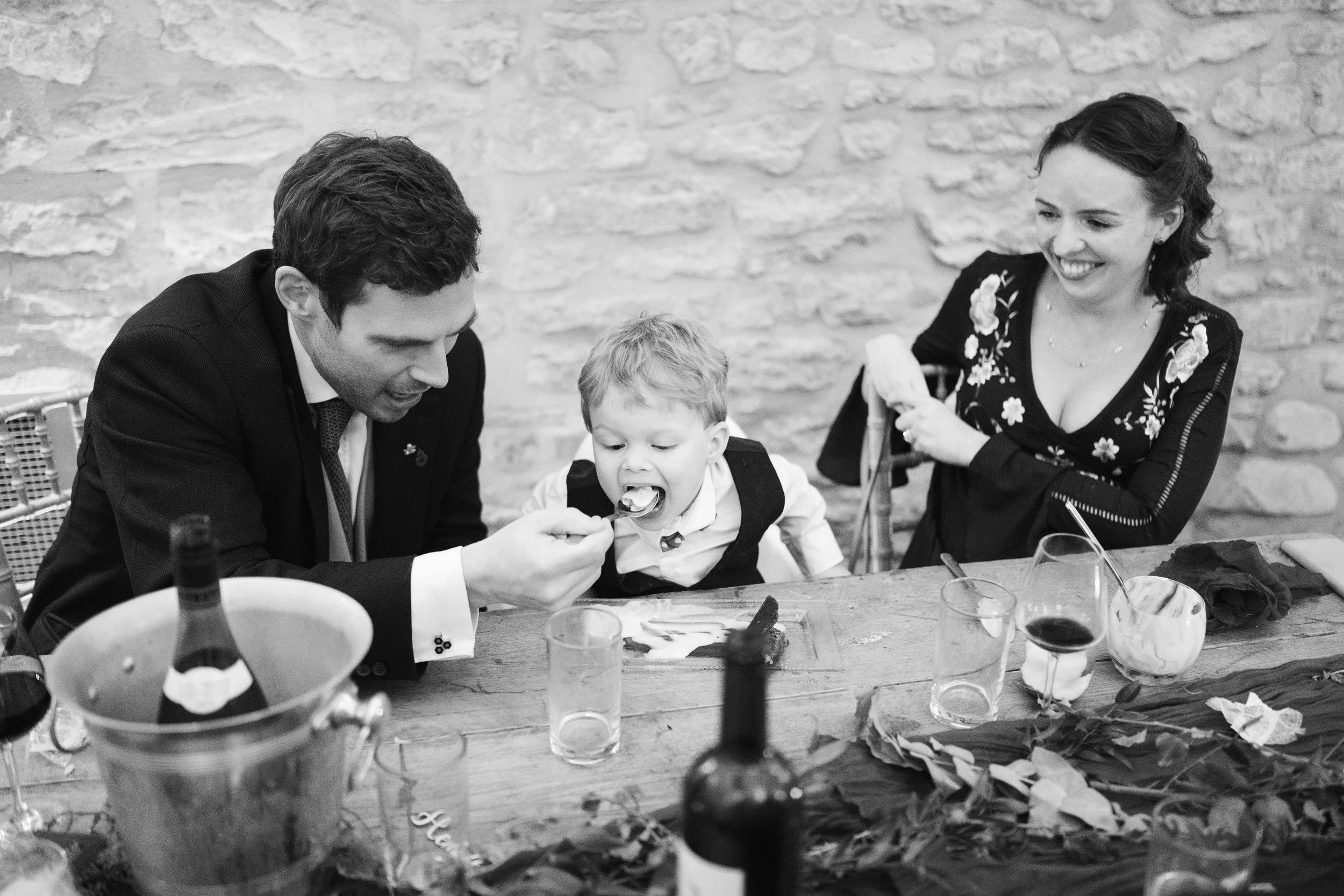 A black and white photo of a family at a festive gathering. A man in a suit is helping a young boy eat with a spoon, while a woman with curly hair and a floral dress looks on and smiles.