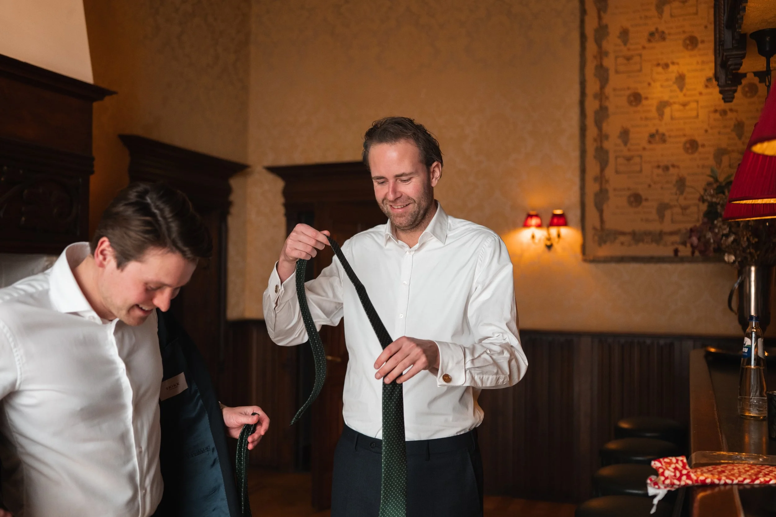 Two men dressed in white shirts smiling and holding ties in a cozy, warmly lit room with wood-paneled walls and decorative lamps.