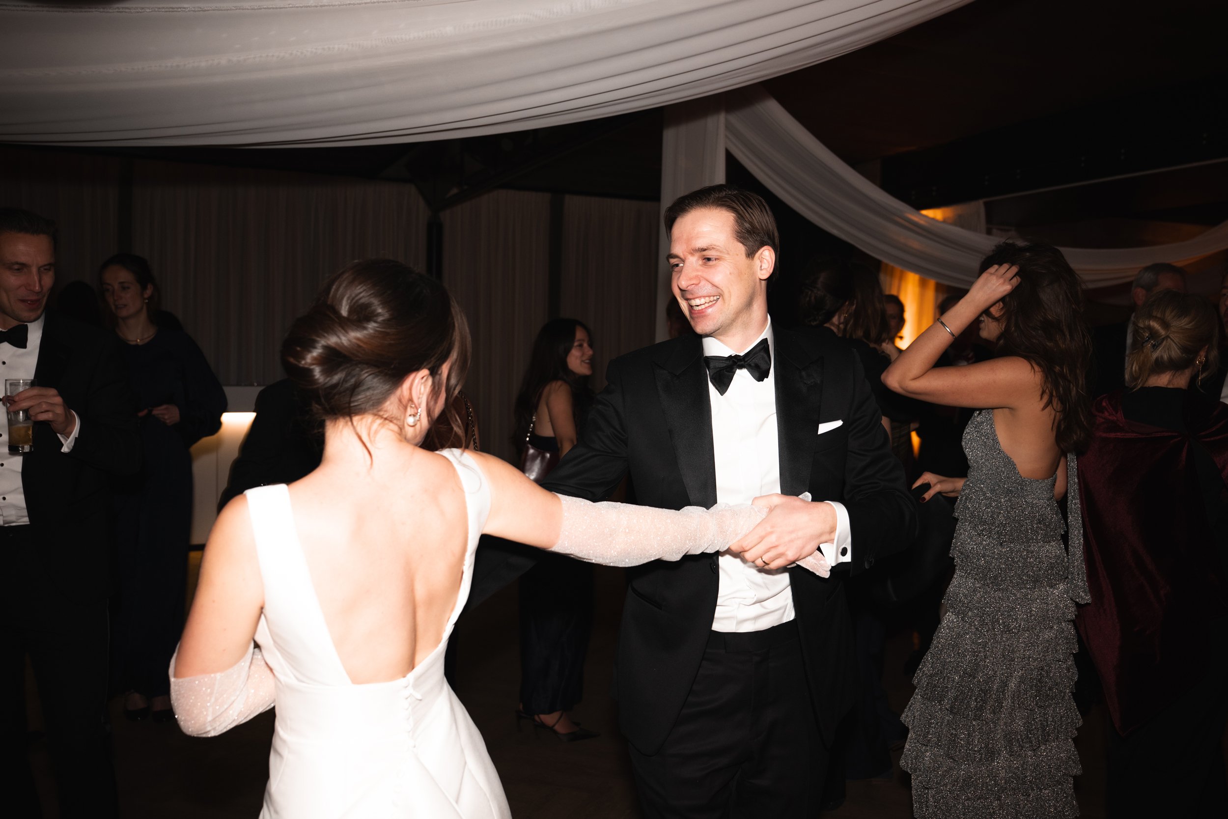 People dancing and socializing at a formal event or wedding reception, with a woman in a white gown and a man in a black tuxedo dancing together in the center.