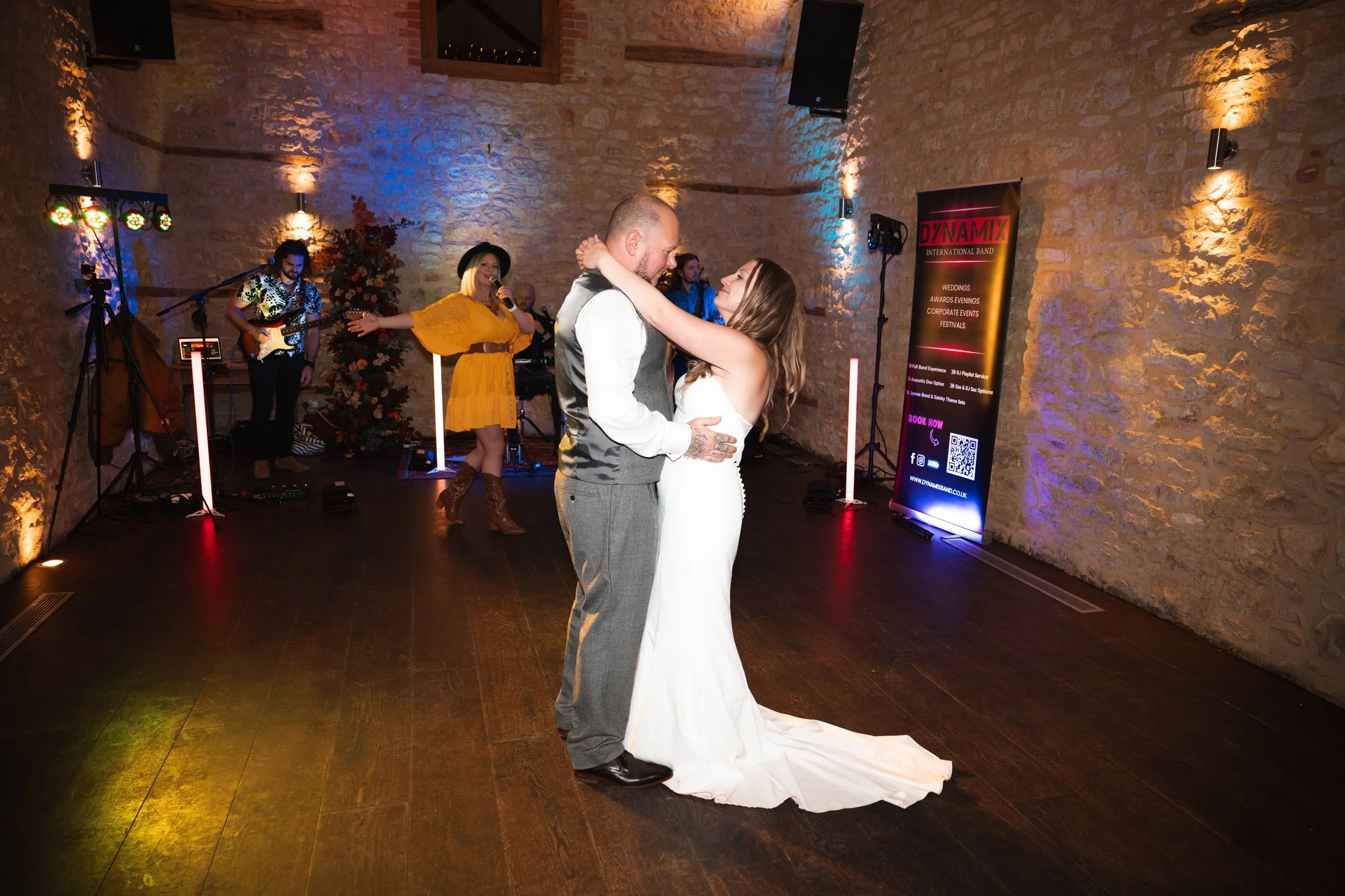 A couple in wedding attire dancing closely in a rustic venue with stone walls. The groom is in a suit vest and trousers, and the bride is in a white dress. A band performs in the background with a woman singing and a guitarist. Colorful lights illumi