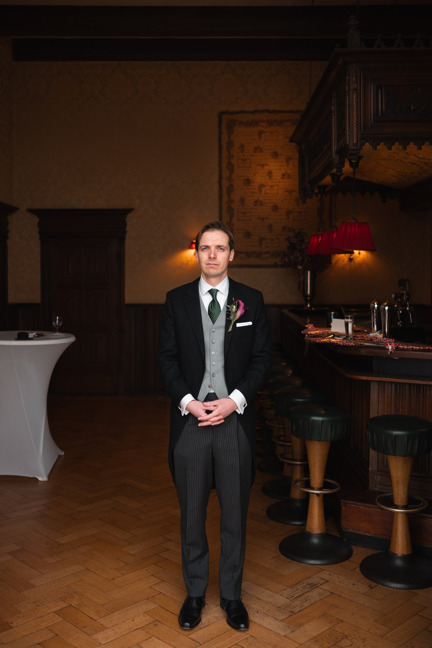 A man dressed in a tuxedo with a boutonniere stands in a dimly lit bar or restaurant, with wooden bar stools and a high table in the background.