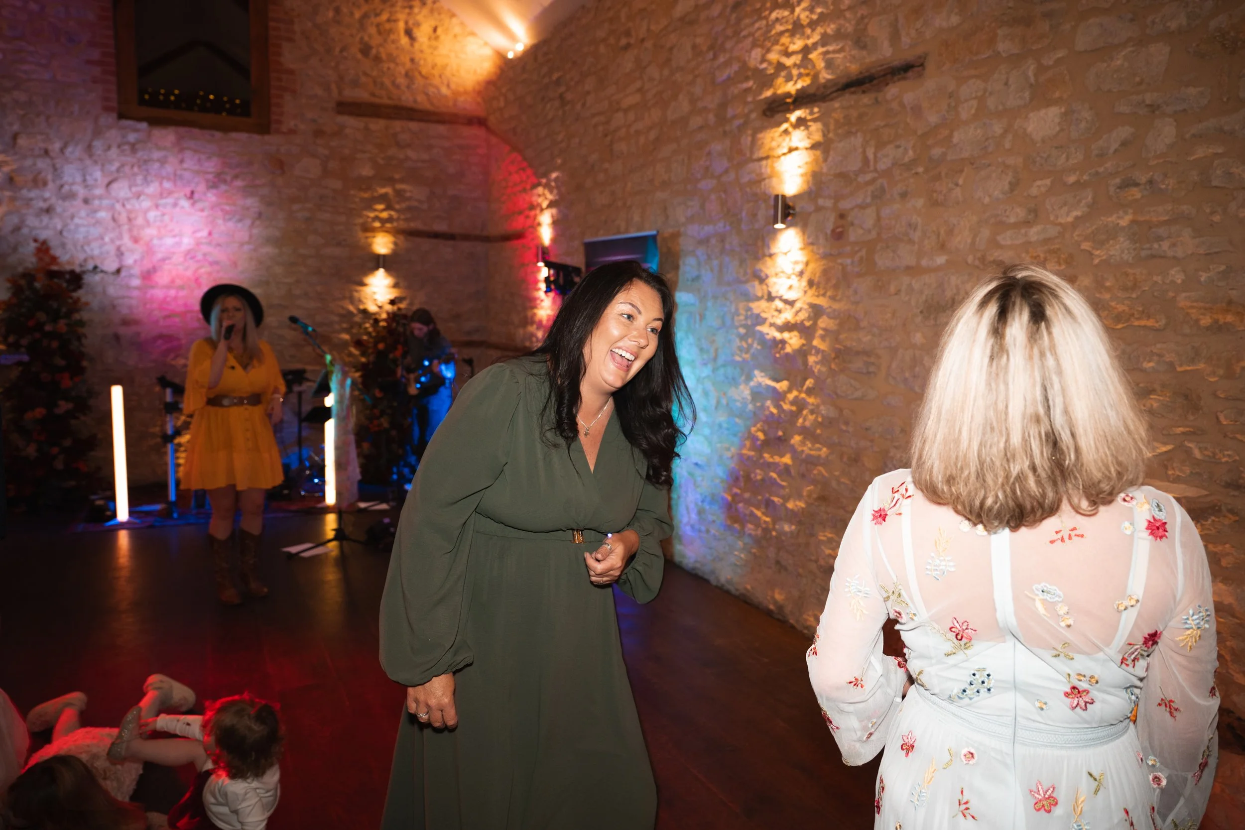Two women are in focus, engaging in a conversation, with a live band performing in colorful stage lighting in the background. One woman has dark hair, is wearing a green dress, and is smiling, while the other has blonde hair, is in a white dress with