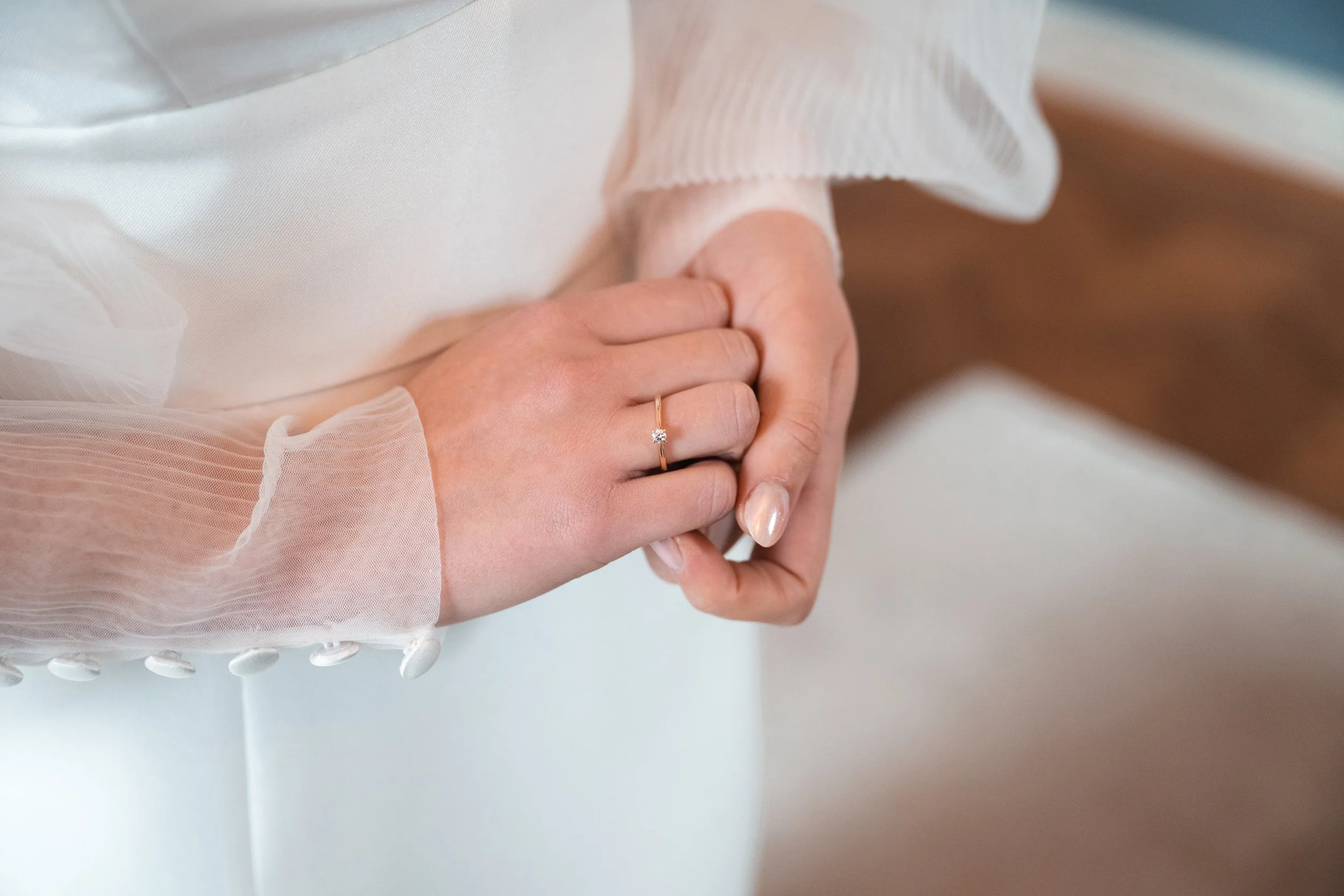 Close-up of a person wearing a white dress with sheer sleeves, holding their hands together, showing a ring with a small gemstone on the finger.