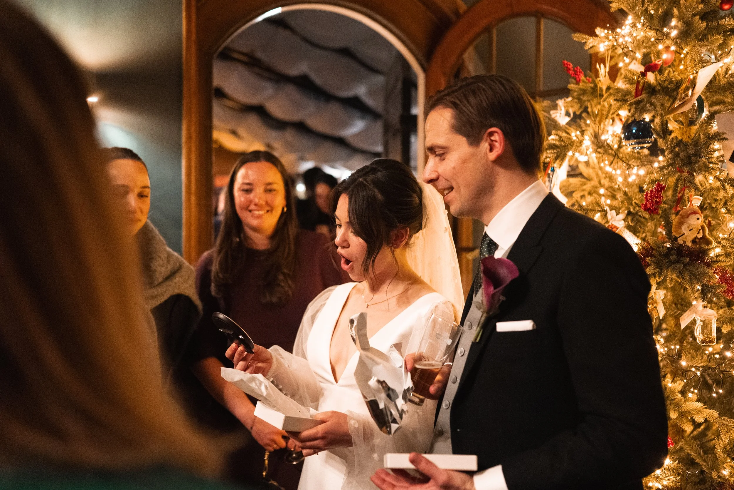 A wedding reception with people gathered around a bride and groom by a decorated Christmas tree, celebrating as the bride reads a card or note.