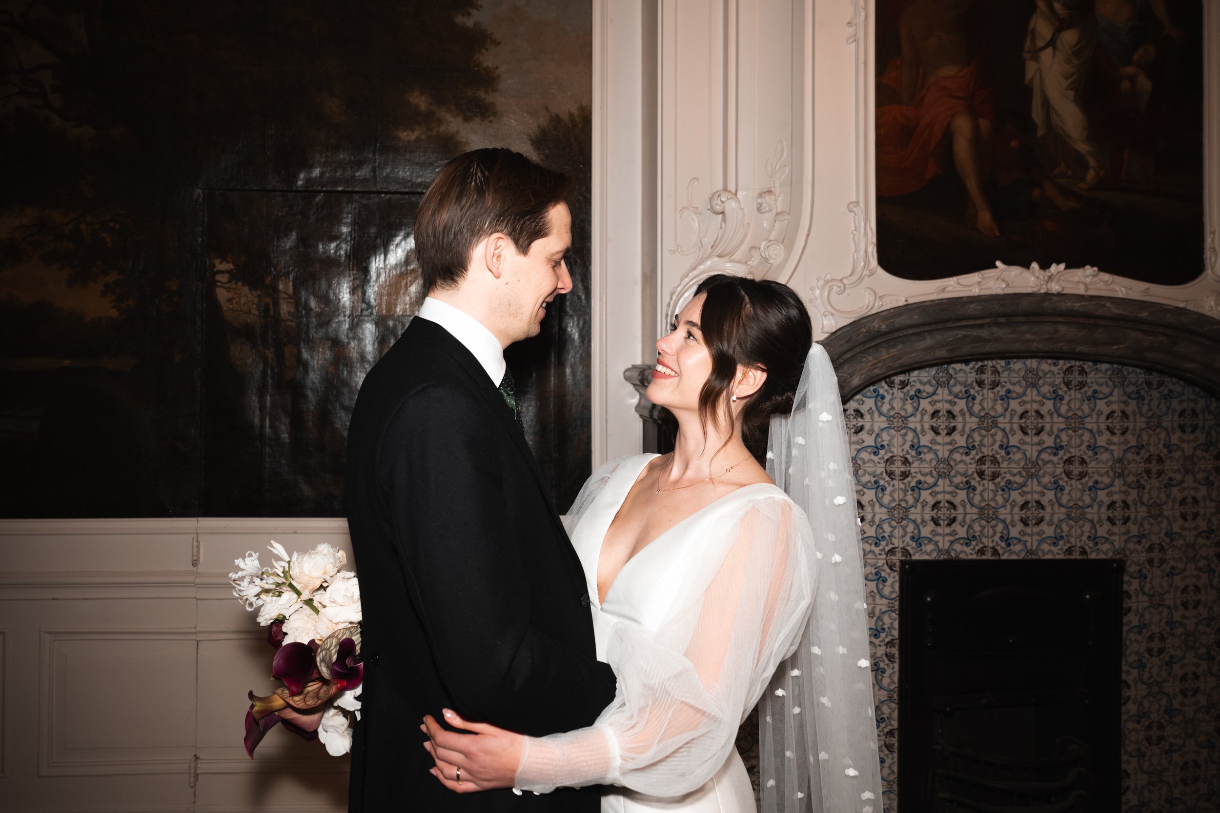 A bride and groom face each other intimately, with the bride smiling up at the groom, holding a bouquet of flowers, inside a decorated indoor wedding venue.
