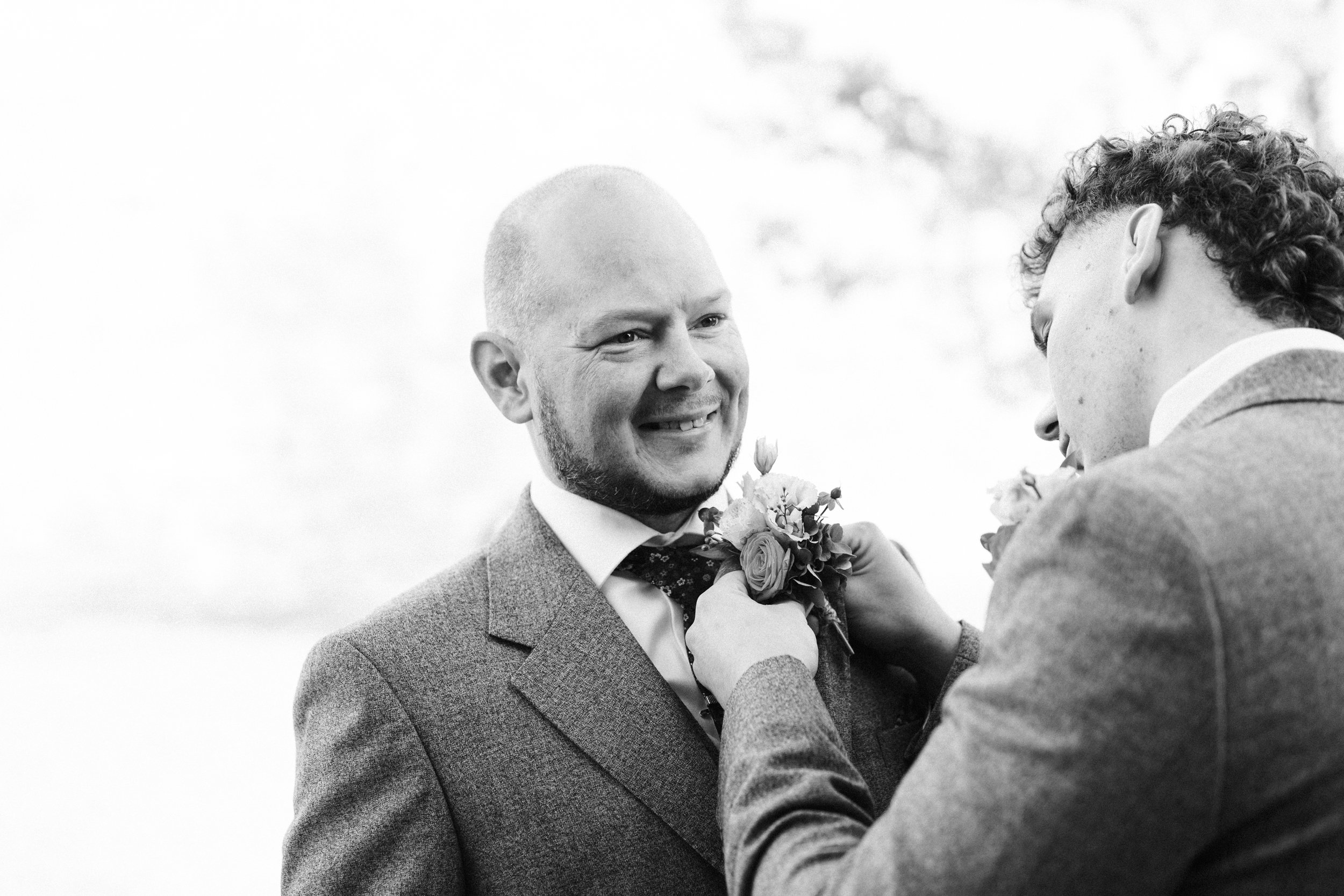 Black and white photo of two men in suits, one placing a boutonniere on the other during a wedding.