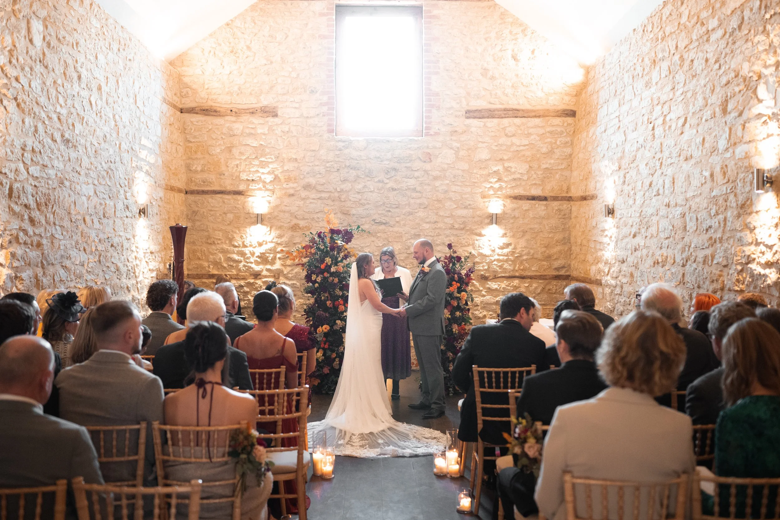 A wedding ceremony inside a rustic stone-walled room with high ceiling, natural light from a window, decorated with flowers and candles, with a bride and groom standing before an officiant.