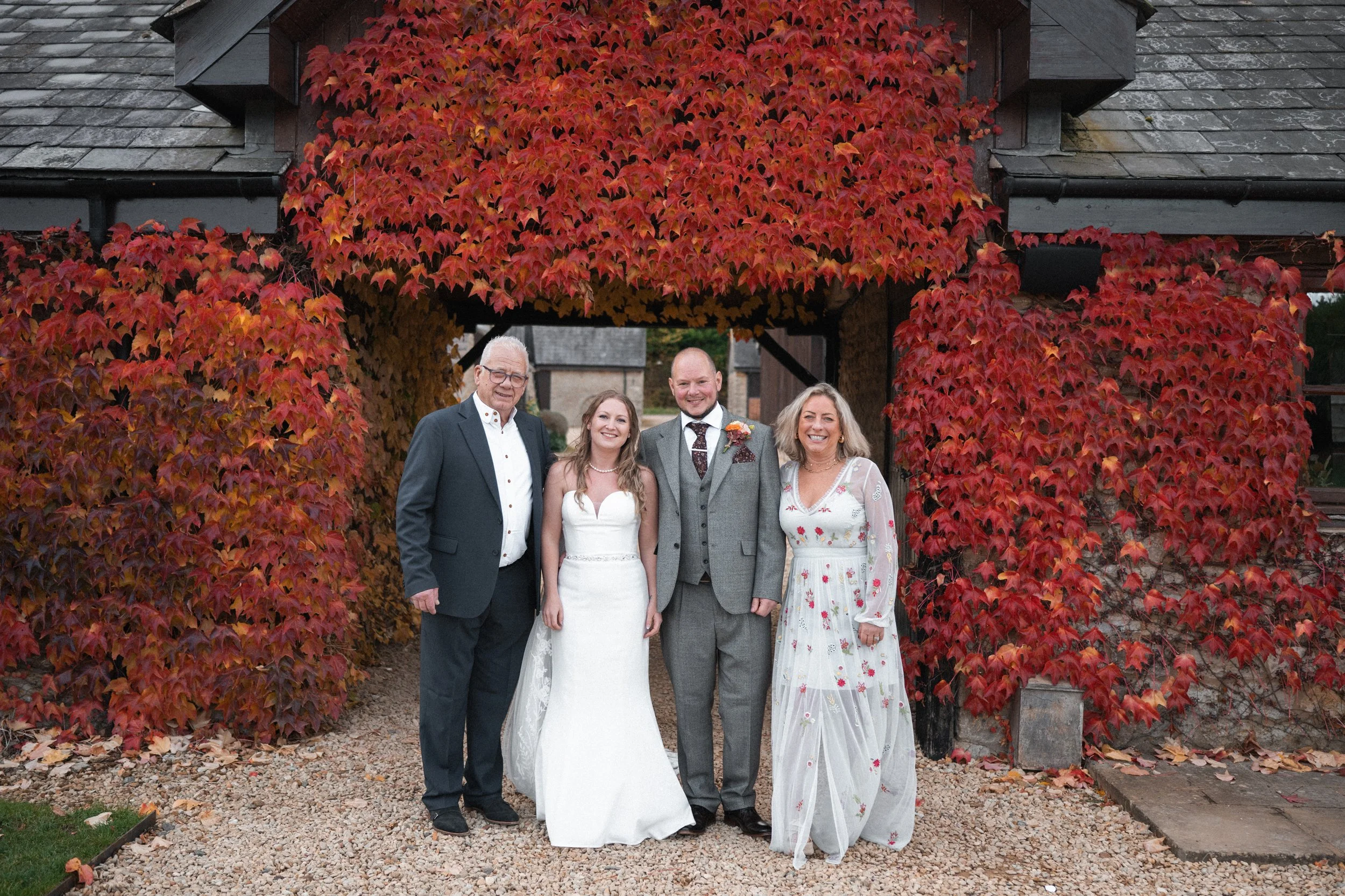 A wedding photo showing four people standing under a wooden archway covered with vibrant red and orange autumn leaves. The group includes a man in a dark suit, a bride in a white wedding dress, a groom in a grey suit, and a woman in a white floral dr