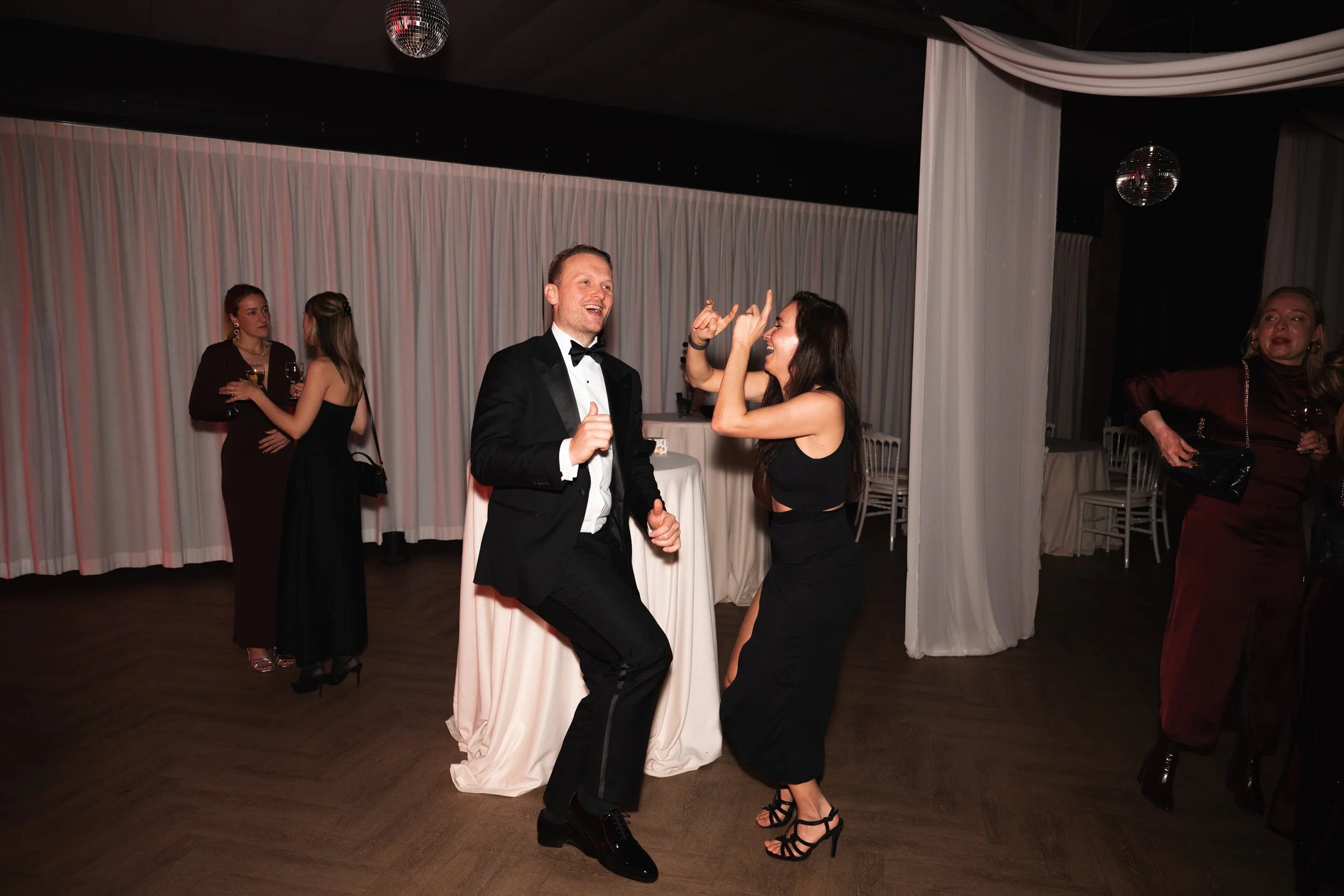 A man in a tuxedo dancing and smiling with a woman in a black dress and high heels at a party or wedding reception. Other people are in the background, talking and holding drinks.