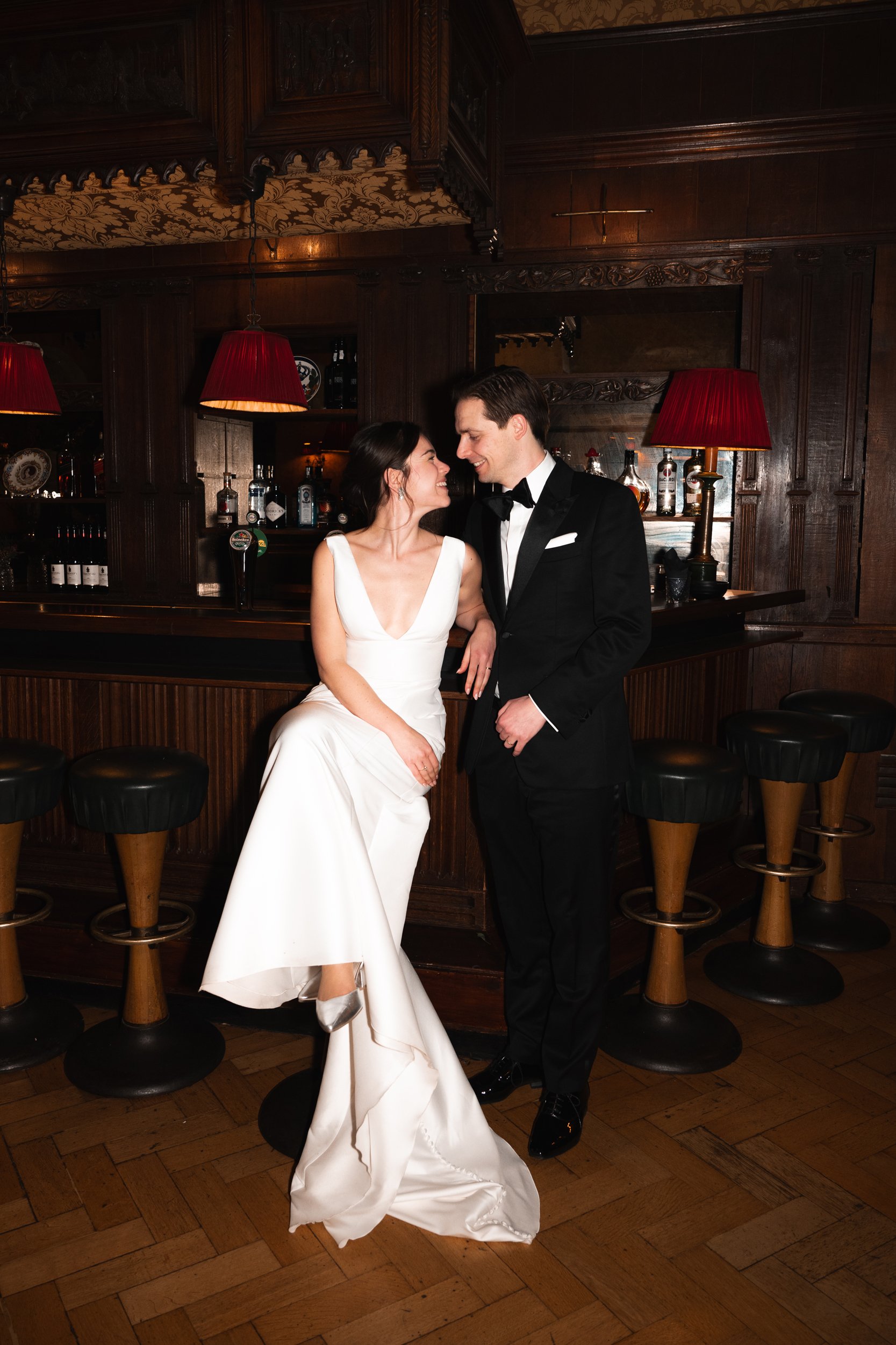 A couple dressed in wedding attire standing by a bar in a dimly lit, wood-paneled room, smiling and looking at each other.
