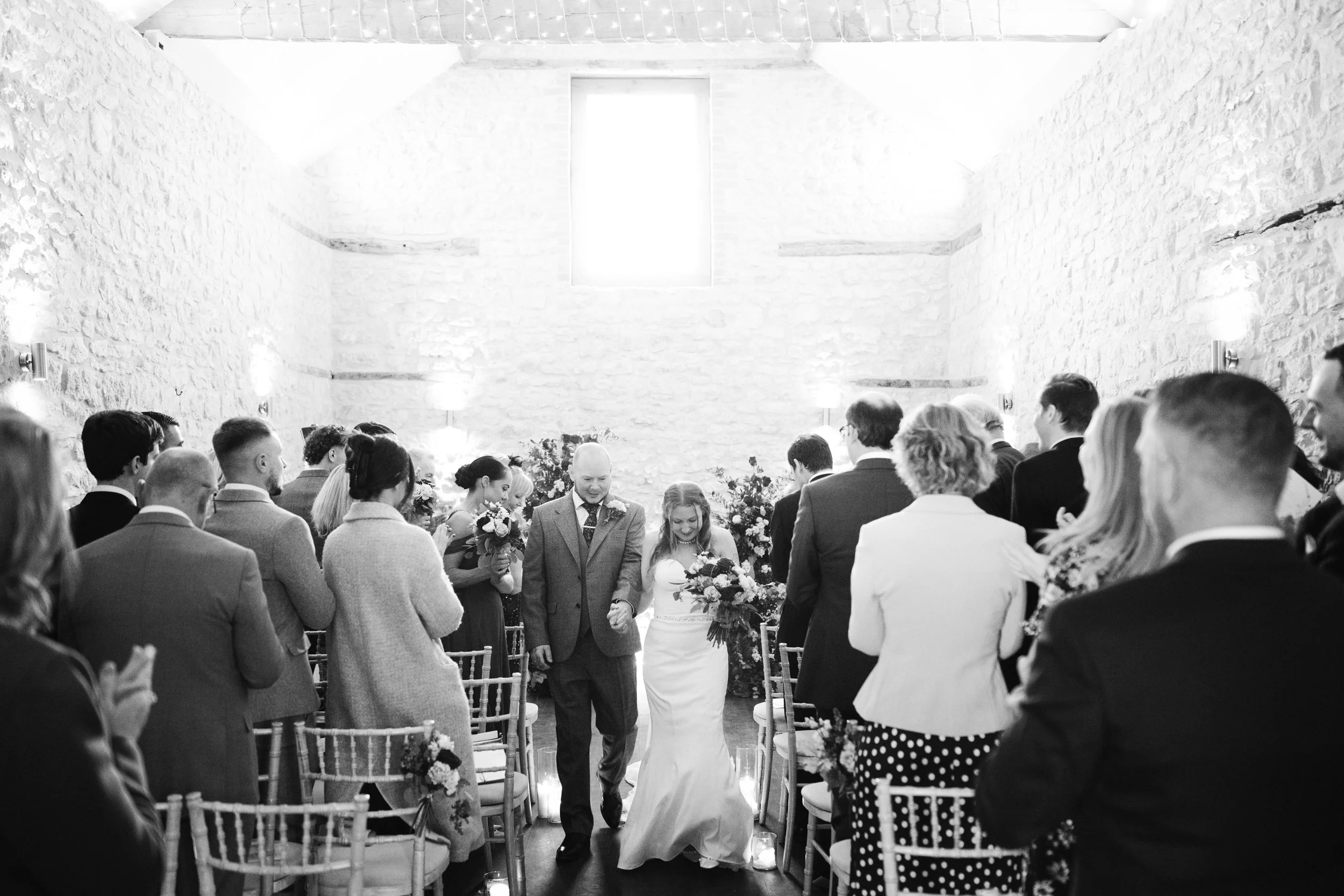 A black and white photo of a wedding ceremony in a rustic stone-walled venue, with the bride walking down the aisle accompanied by an older man, likely her father, surrounded by guests sitting and standing.