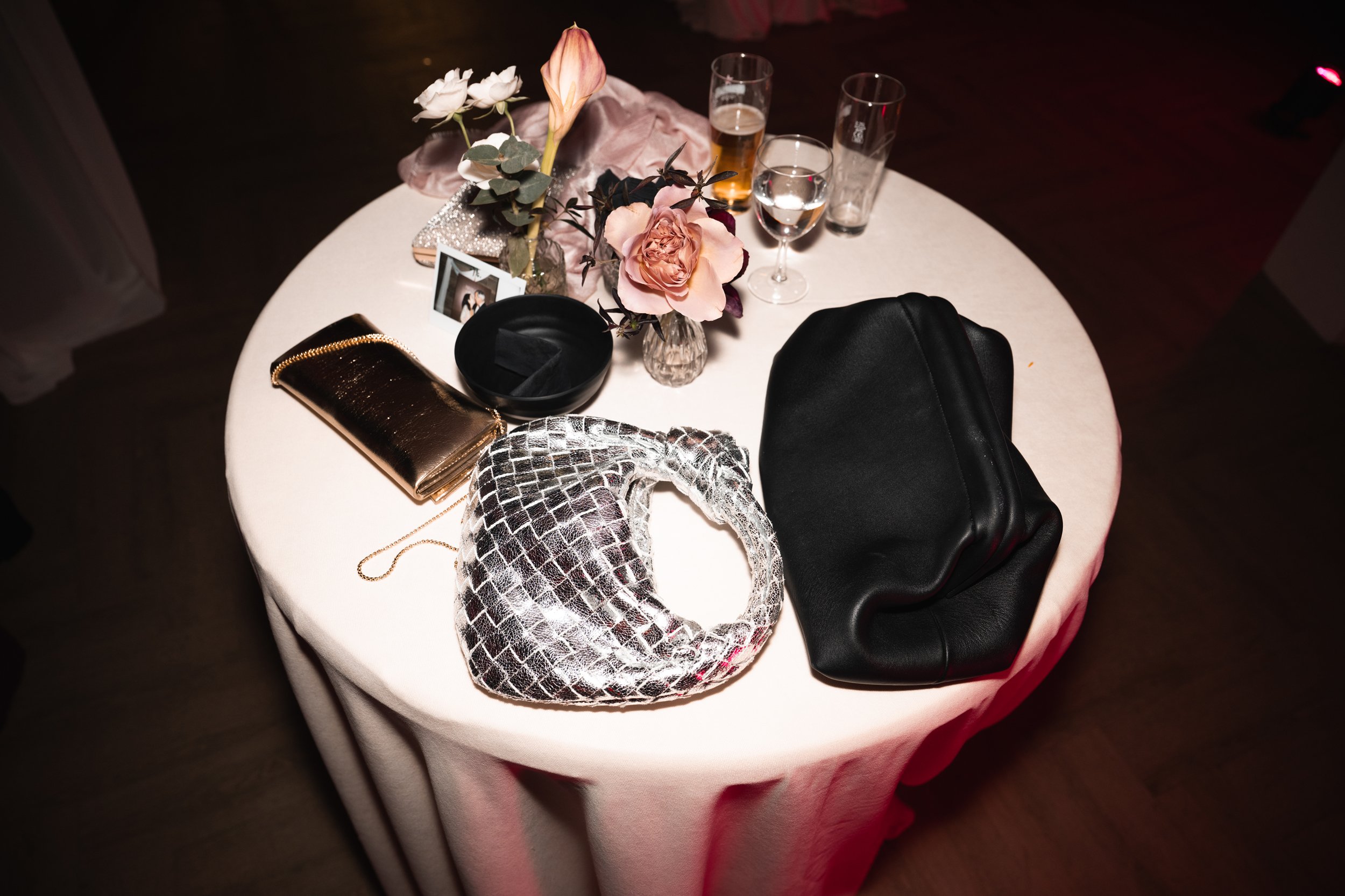 A round table with a white tablecloth holding floral arrangements, glasses with drinks, a gold clutch, a black pouch, a black hat, and a silver-patterned clutch, with a small framed photograph in a dimly lit setting.