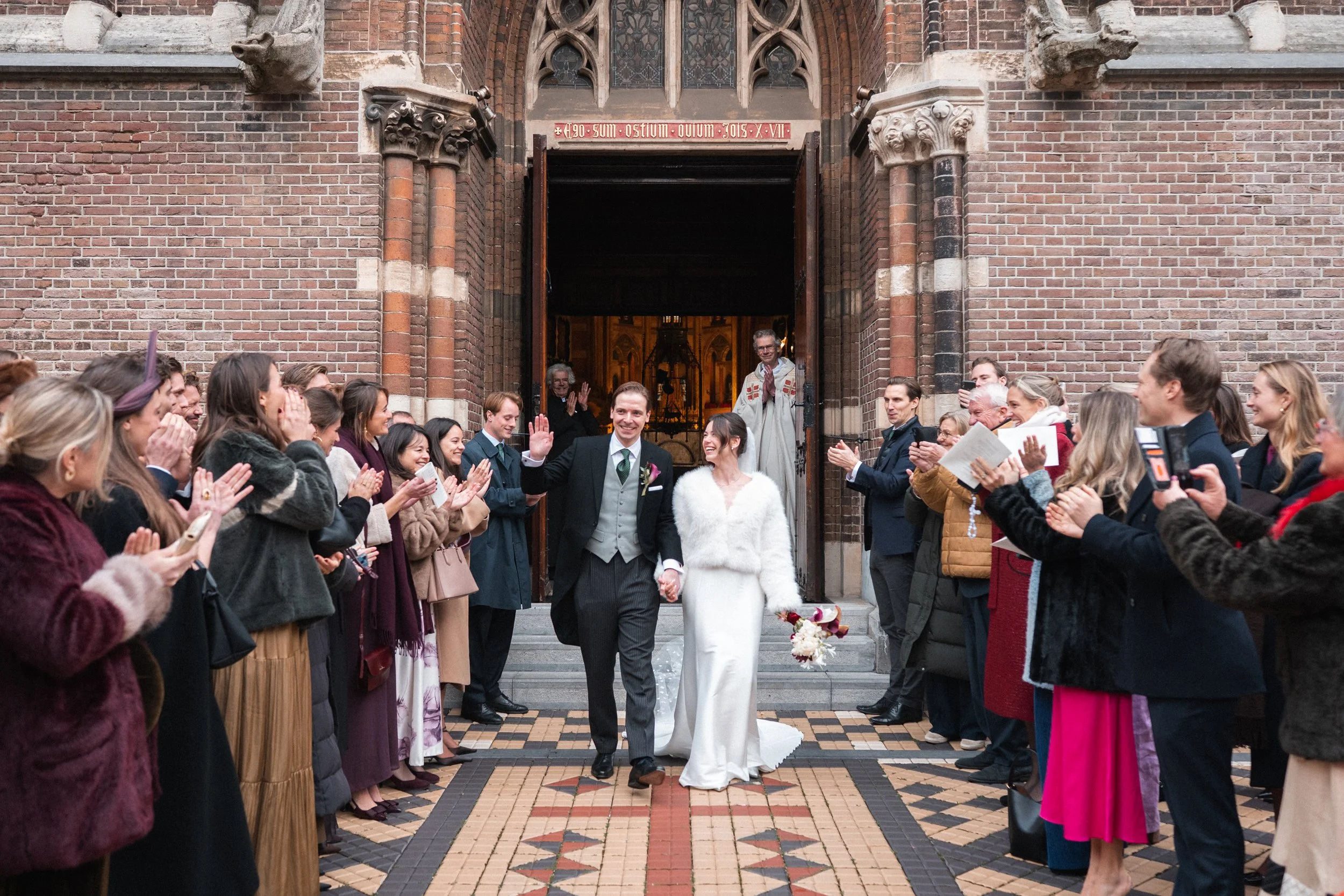 A newlywed couple walking out of a church while friends and family celebrate and clap.