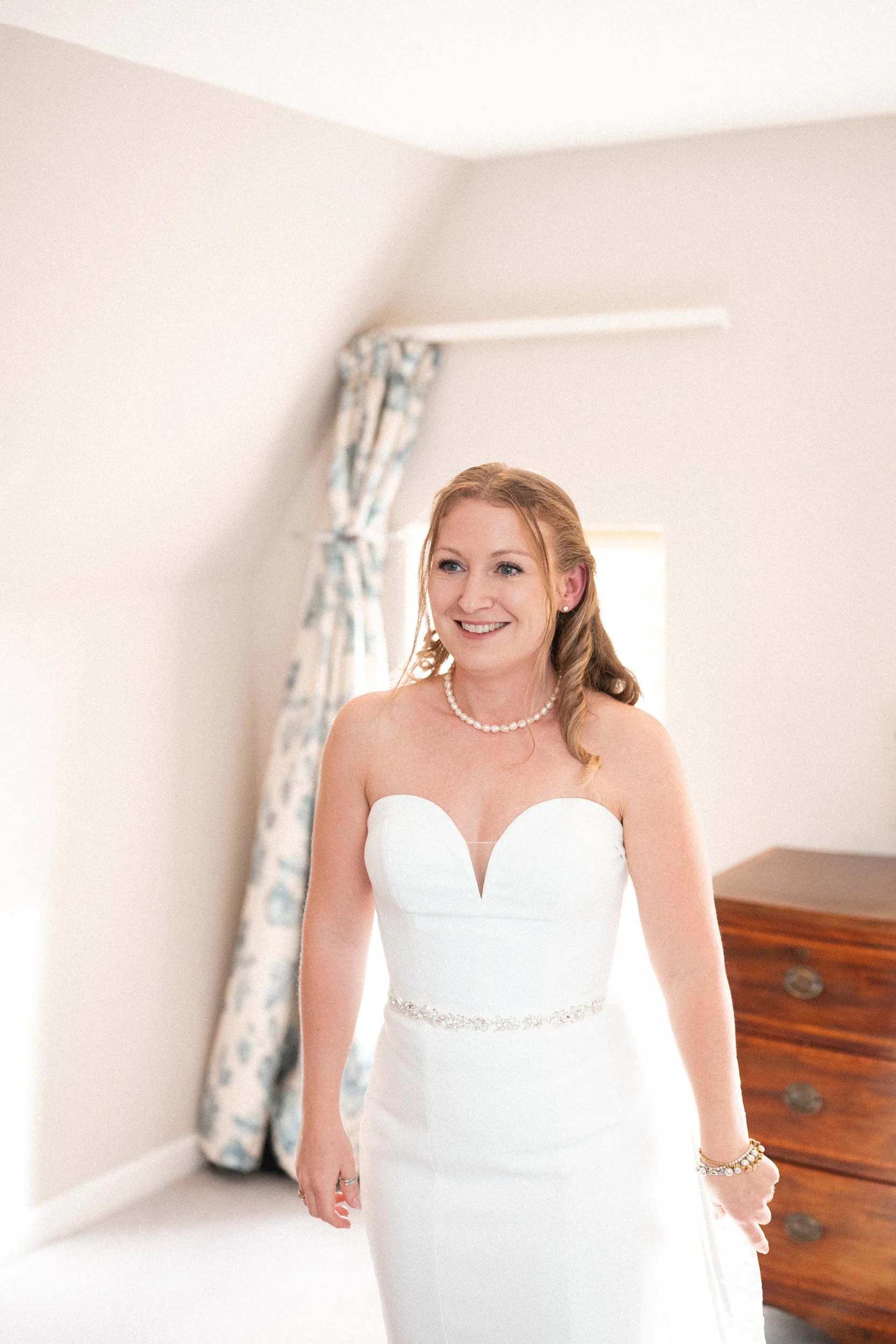 A smiling woman in a white wedding dress standing in a bright room with curtains and a dresser.
