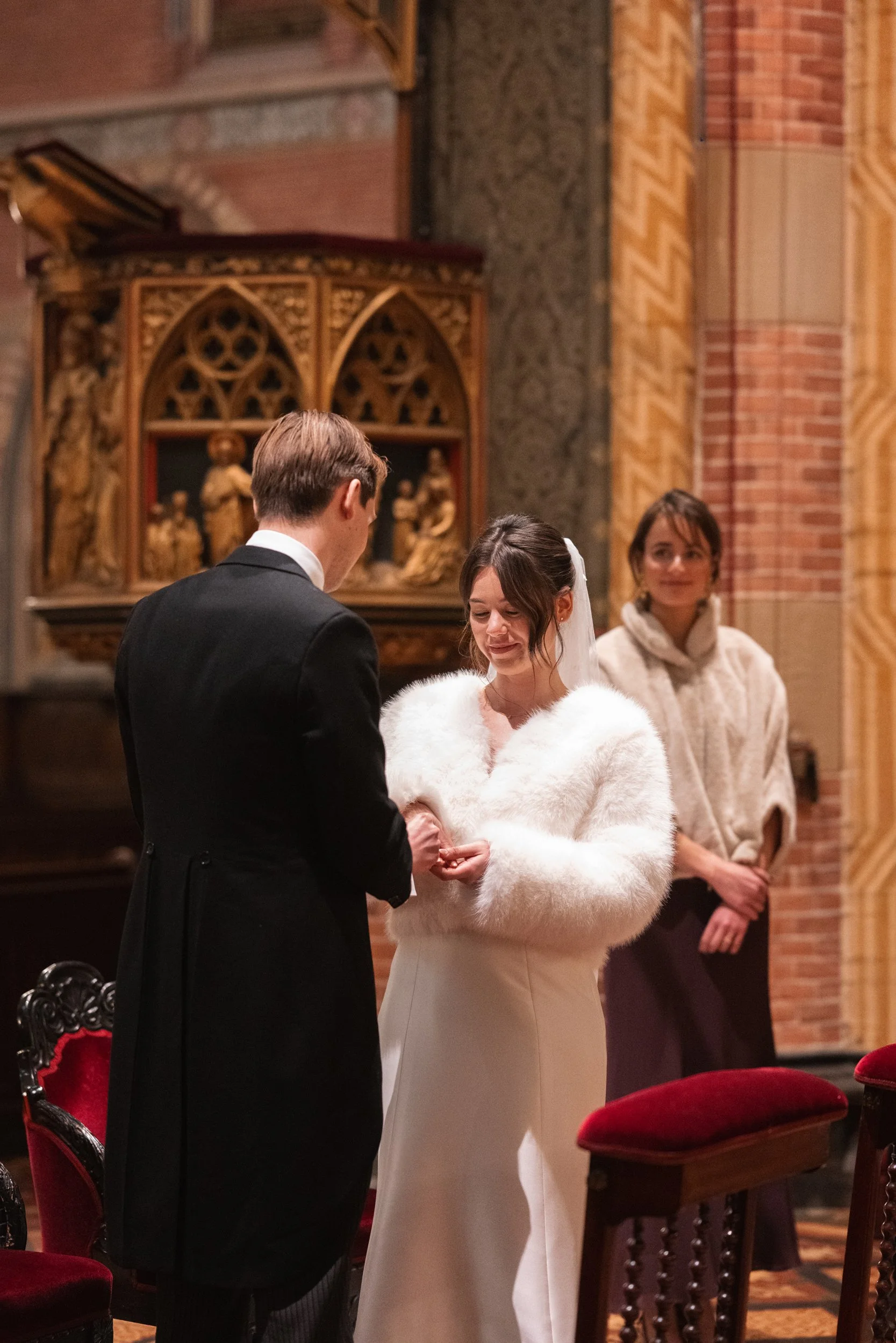 A wedding ceremony taking place in a church with a bride and groom exchanging rings, and a woman in the background watching.